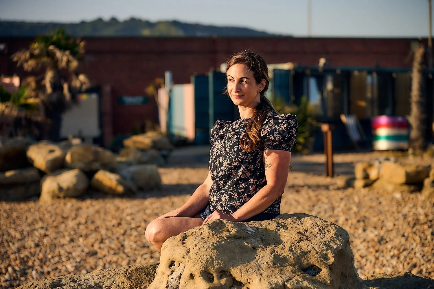 A woman with long hair sitting on a large rock at a beach during sunset, wearing a floral blouse, with rocks and beach in the background.