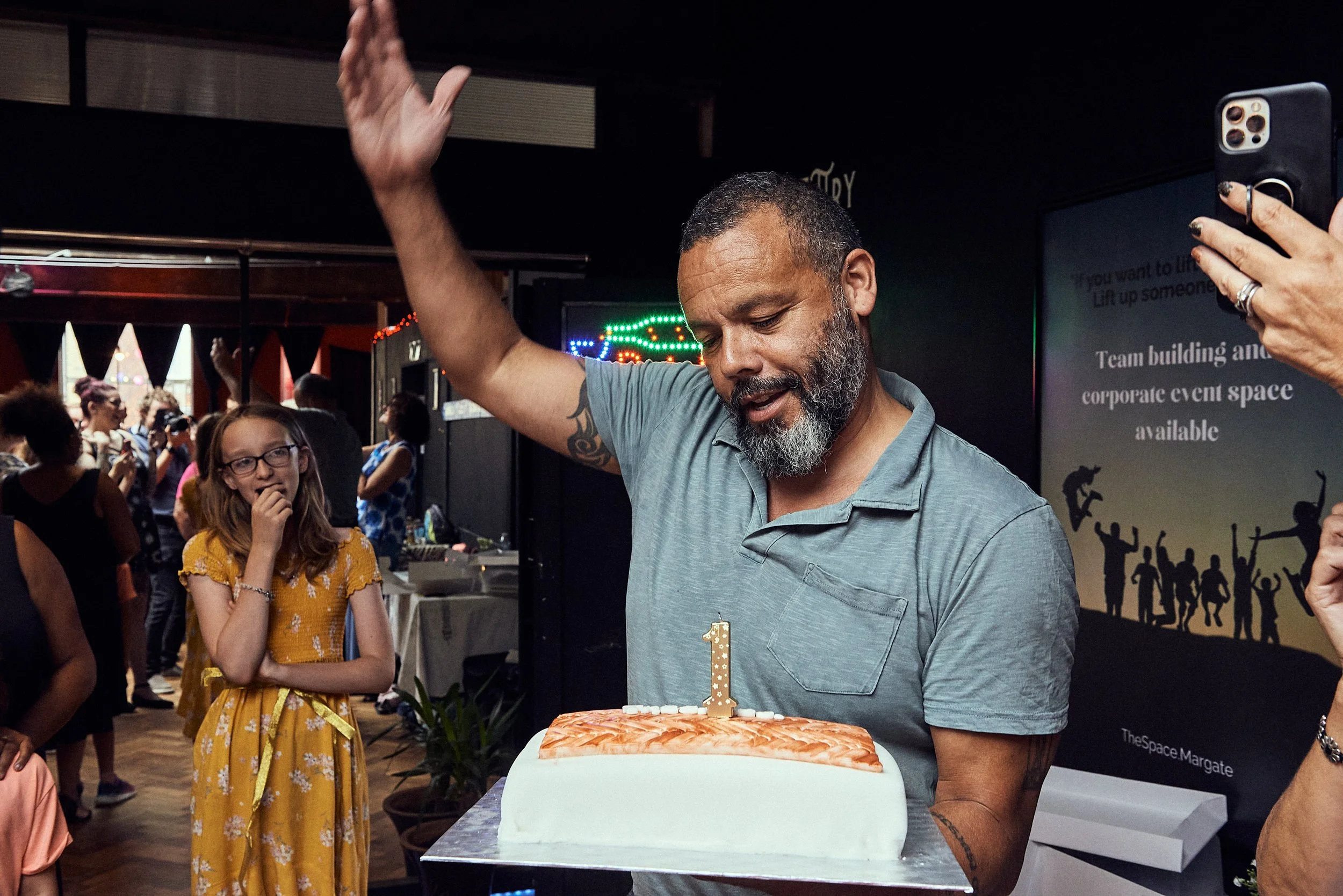 A man with a beard and gray hair holding a tray with a cake topped with a single lit candle, surrounded by a crowd in a celebration setting, possibly a birthday, with a woman in glasses and a yellow dress nearby.