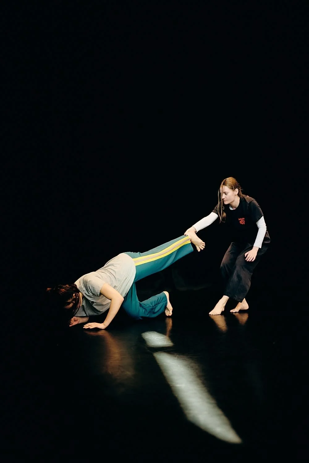 Two women practicing a physical exercise or dance move in a dark studio, one woman is kneeling and holding the other woman by the ankles as she leans forward with her head on the floor.