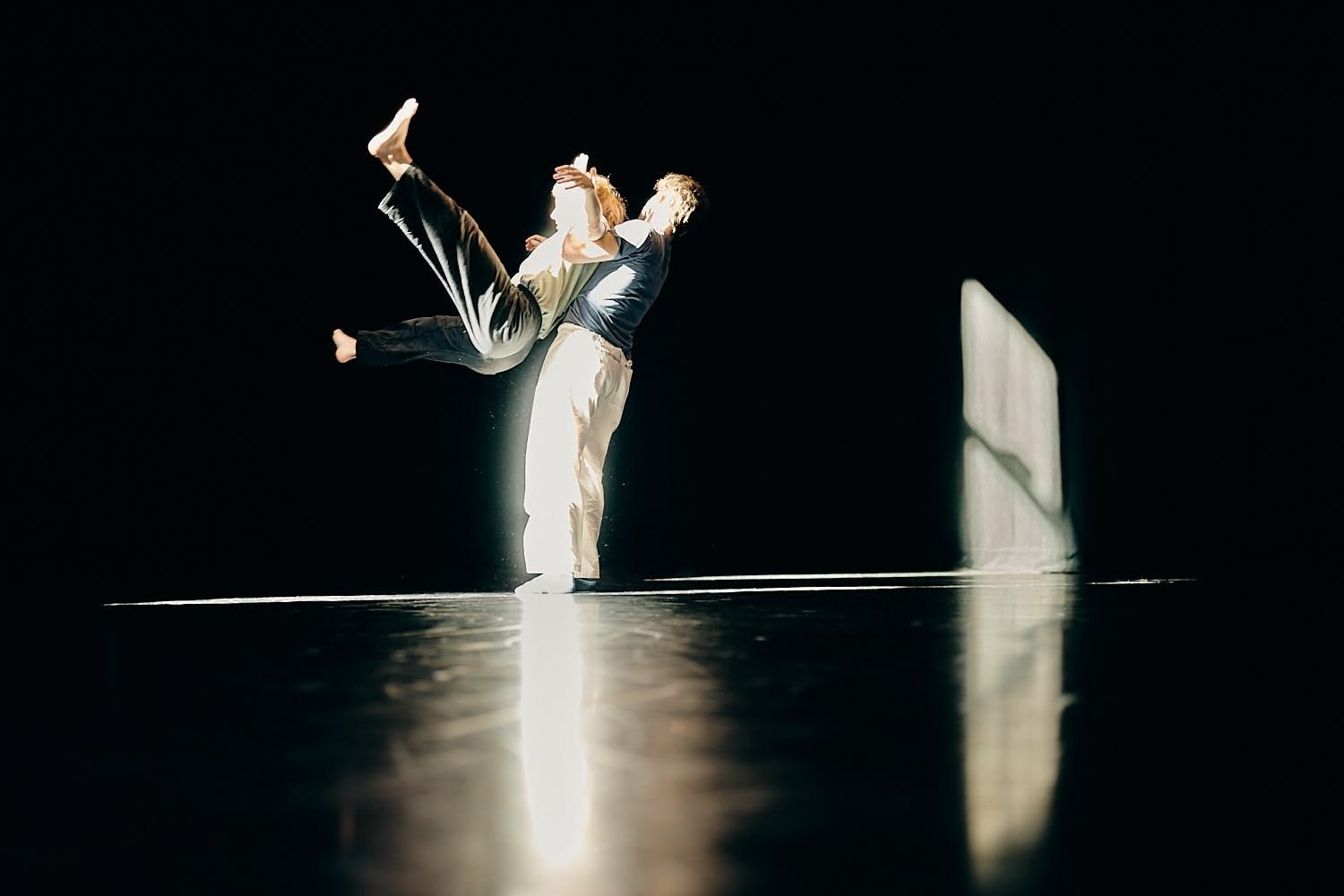 Two people dancing in a dark studio, with sunlight streaming through a window creating a bright spot on the floor.