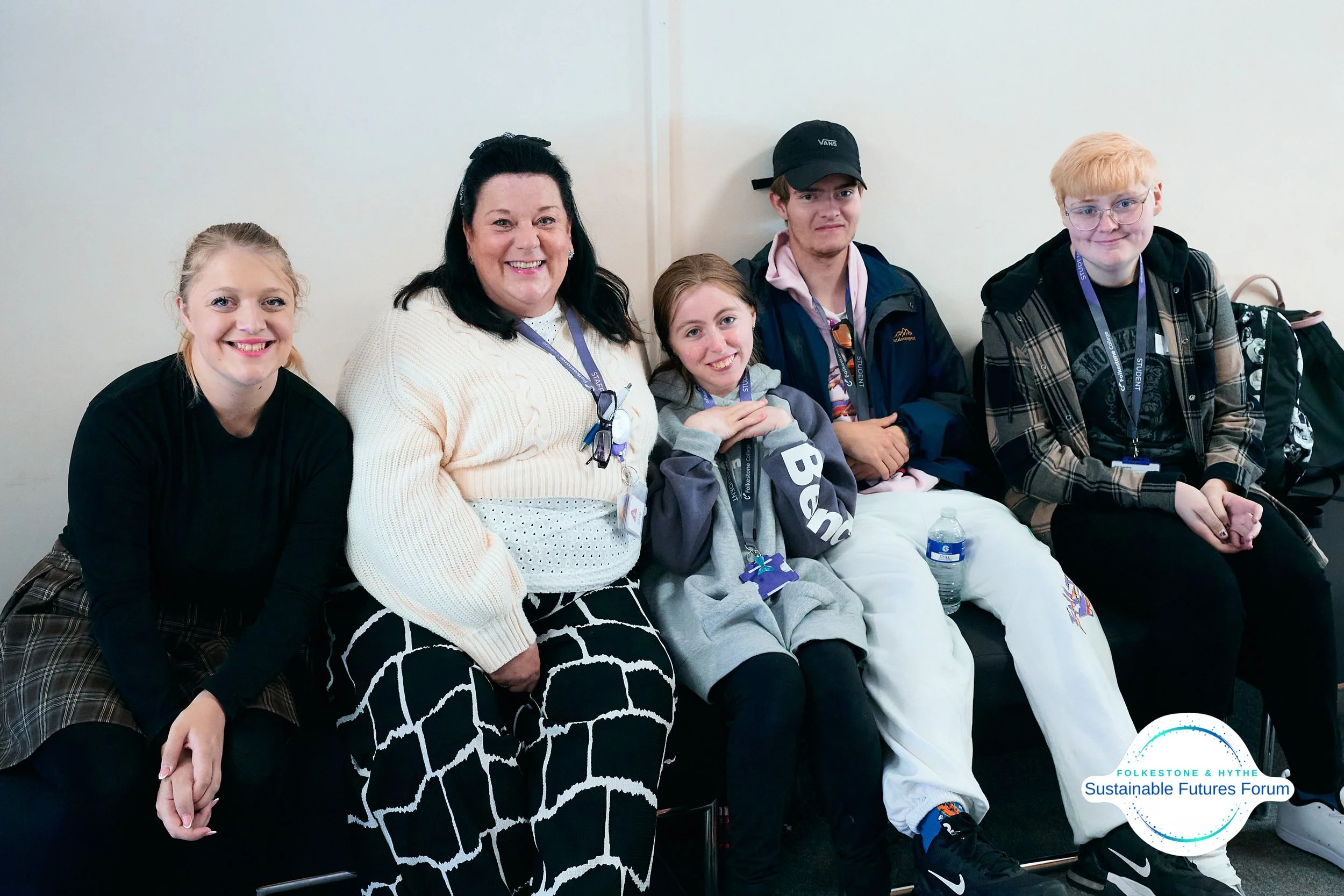 Five smiling people, including women and teenagers, sitting on a bench, wearing casual clothing, at a forum or conference.