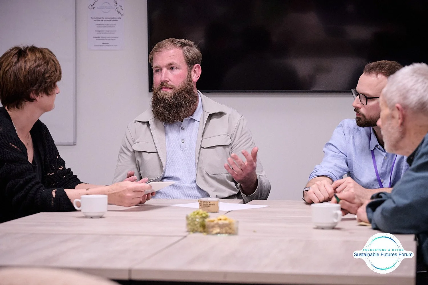 Four people engaged in a discussion at a meeting, sitting around a table with coffee cups and desserts, in a conference room.