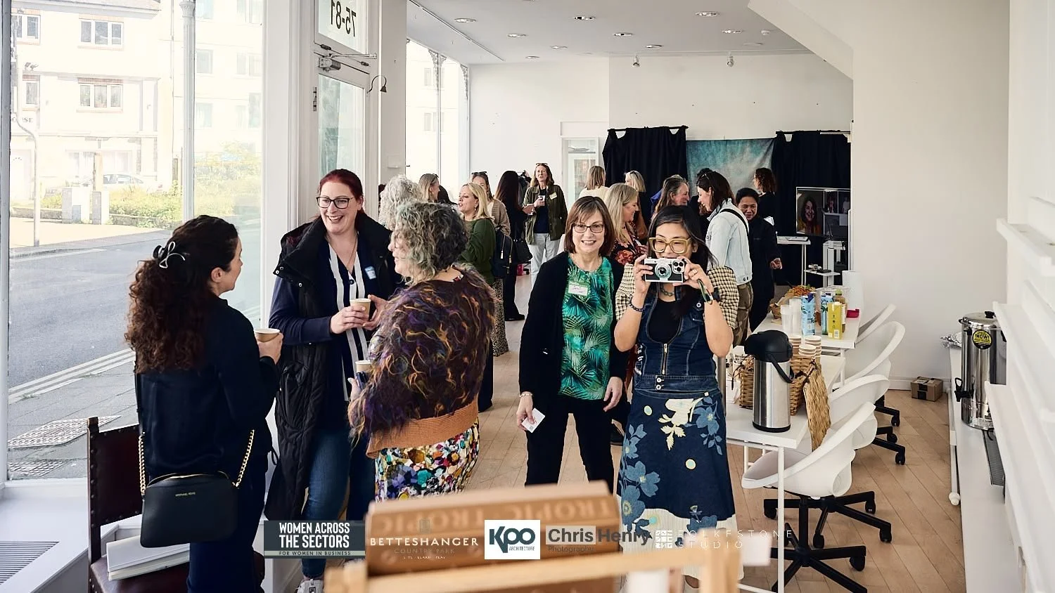 Women at a networking event chatting and taking photos in a bright, modern space with large windows.
