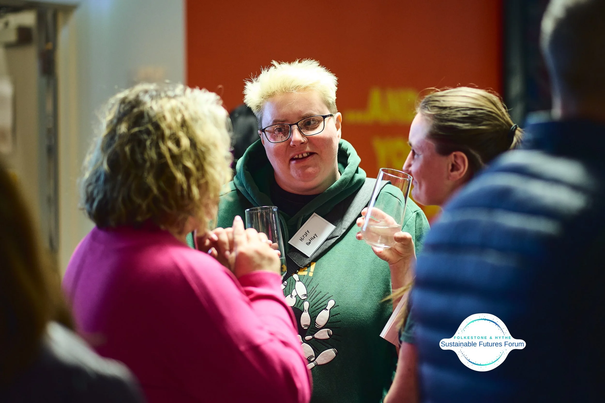 People engaged in conversation at the Folkesone & Hythe Sustainable Futures Forum, with one woman wearing glasses and a green hoodie, holding a glass, and a woman in a pink jacket, and another woman in a blue jacket nearby.
