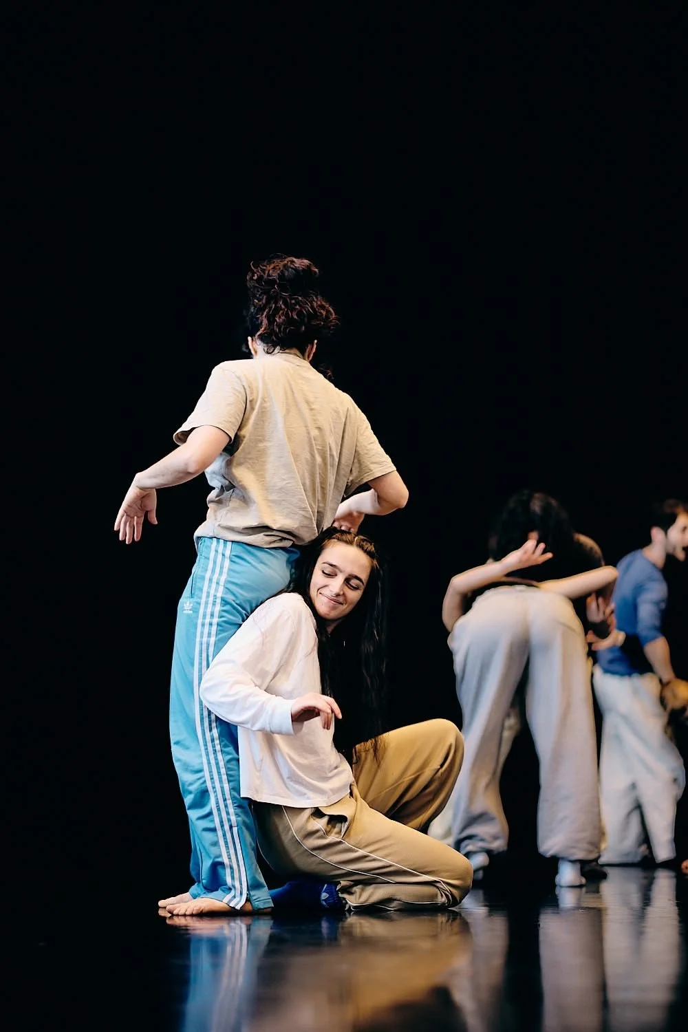 Dancers rehearsing on stage, one dancer smiling with eyes closed, others with backs turned, all in casual dancewear, against a black background.