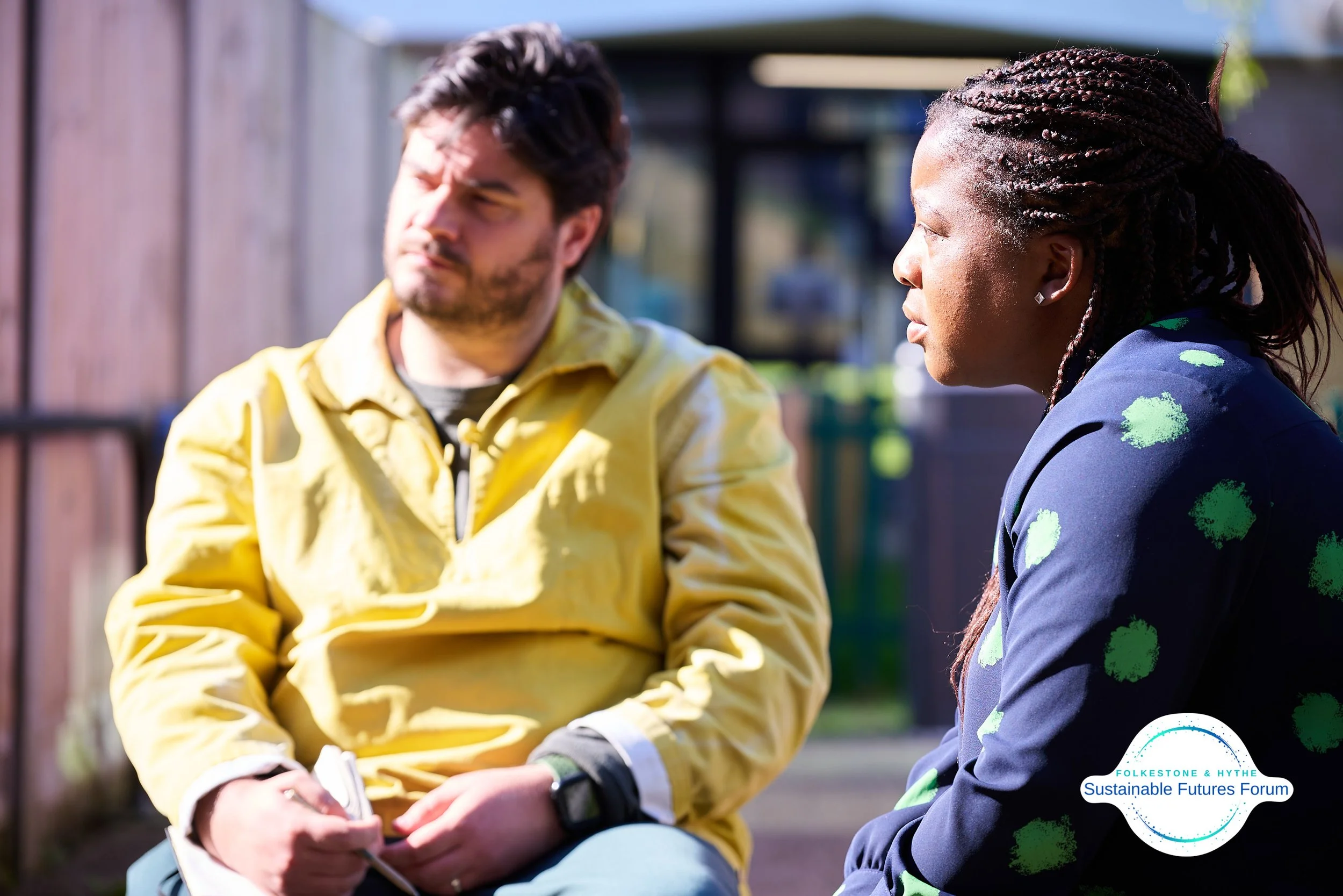 Two people sitting outdoors having a conversation, one wearing a yellow jacket and the other in a dark blazer with green patterns, with a logo that reads 'Folkes tone & Hythe Sustainable Futures Forum' in the bottom right corner.