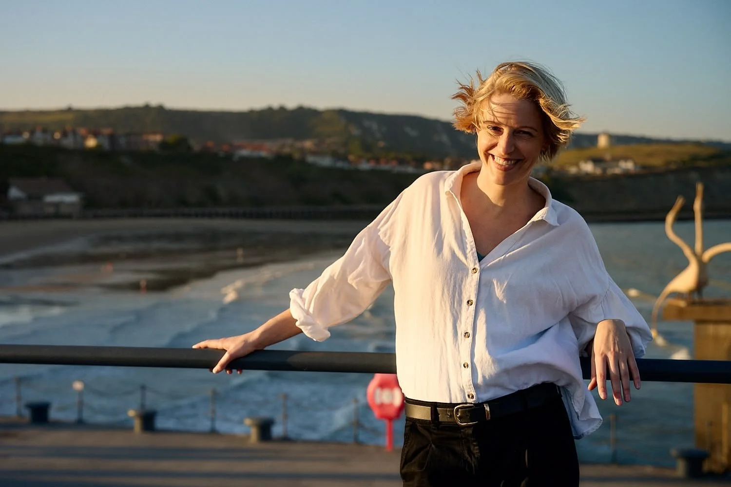 A woman with short blonde hair wearing a white button-up shirt and black pants, smiling and standing outdoors near a body of water with hills and houses in the background.