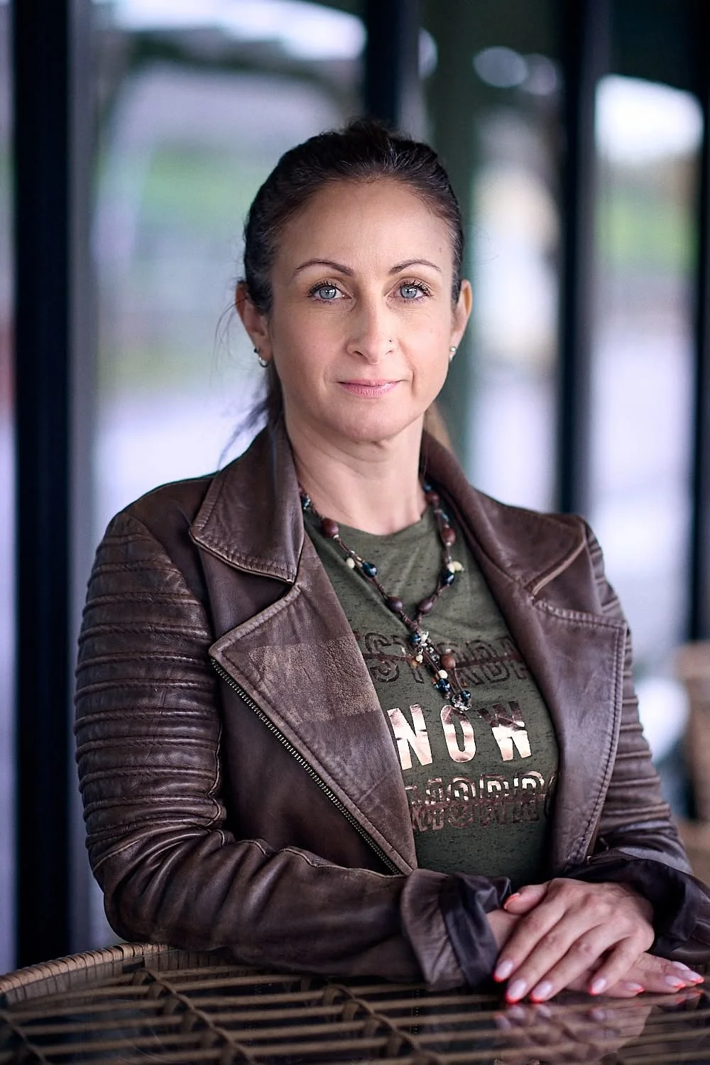 A woman with dark hair pulled back, wearing a brown leather jacket over a green t-shirt with text, and a beaded necklace, standing indoors near windows with natural light.