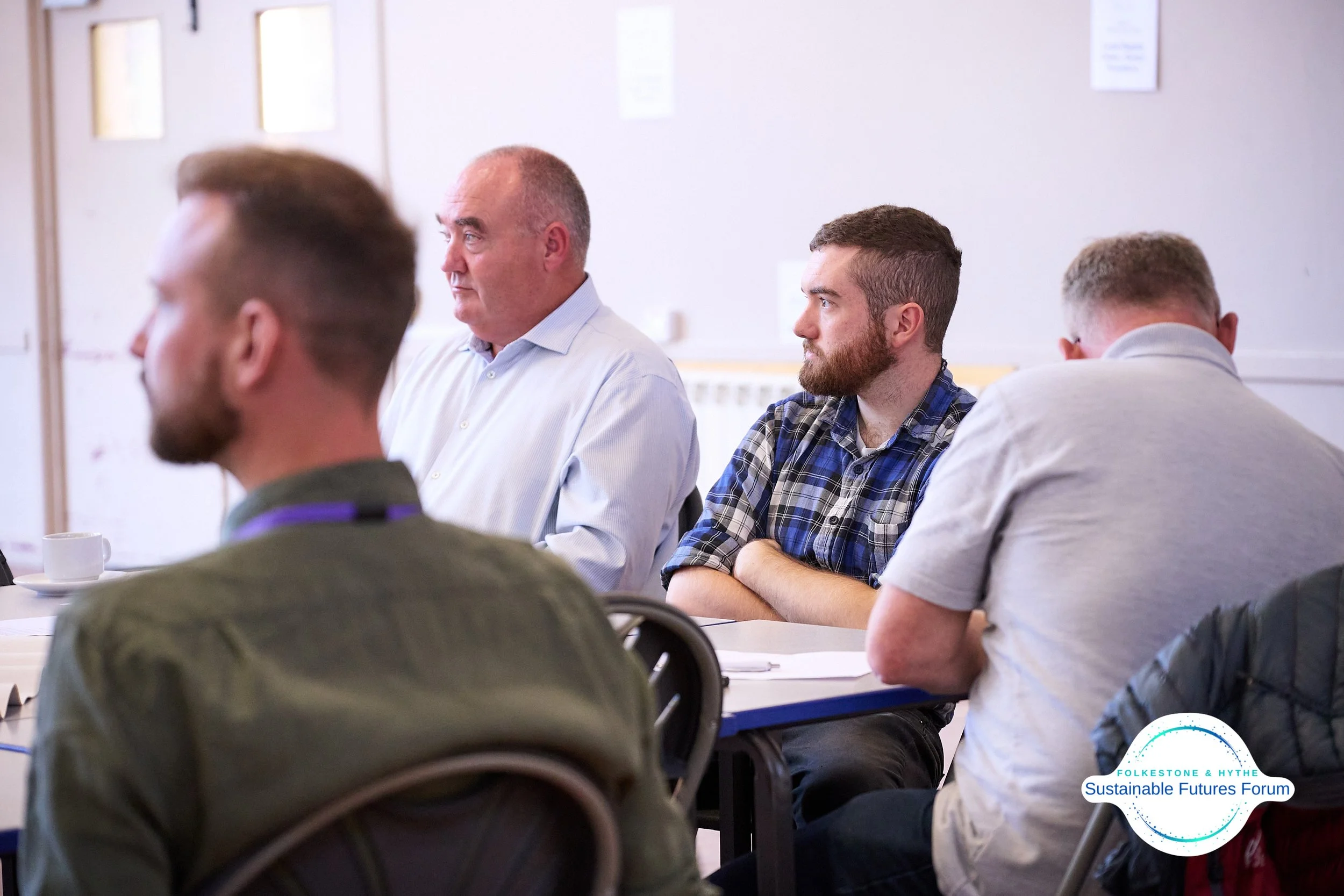 Four men sitting at a table during a forum or meeting, with a whiteboard and papers on the table and a logo that says "Folkes tone & Hythe - Sustainable Futures Forum" in the bottom right corner.