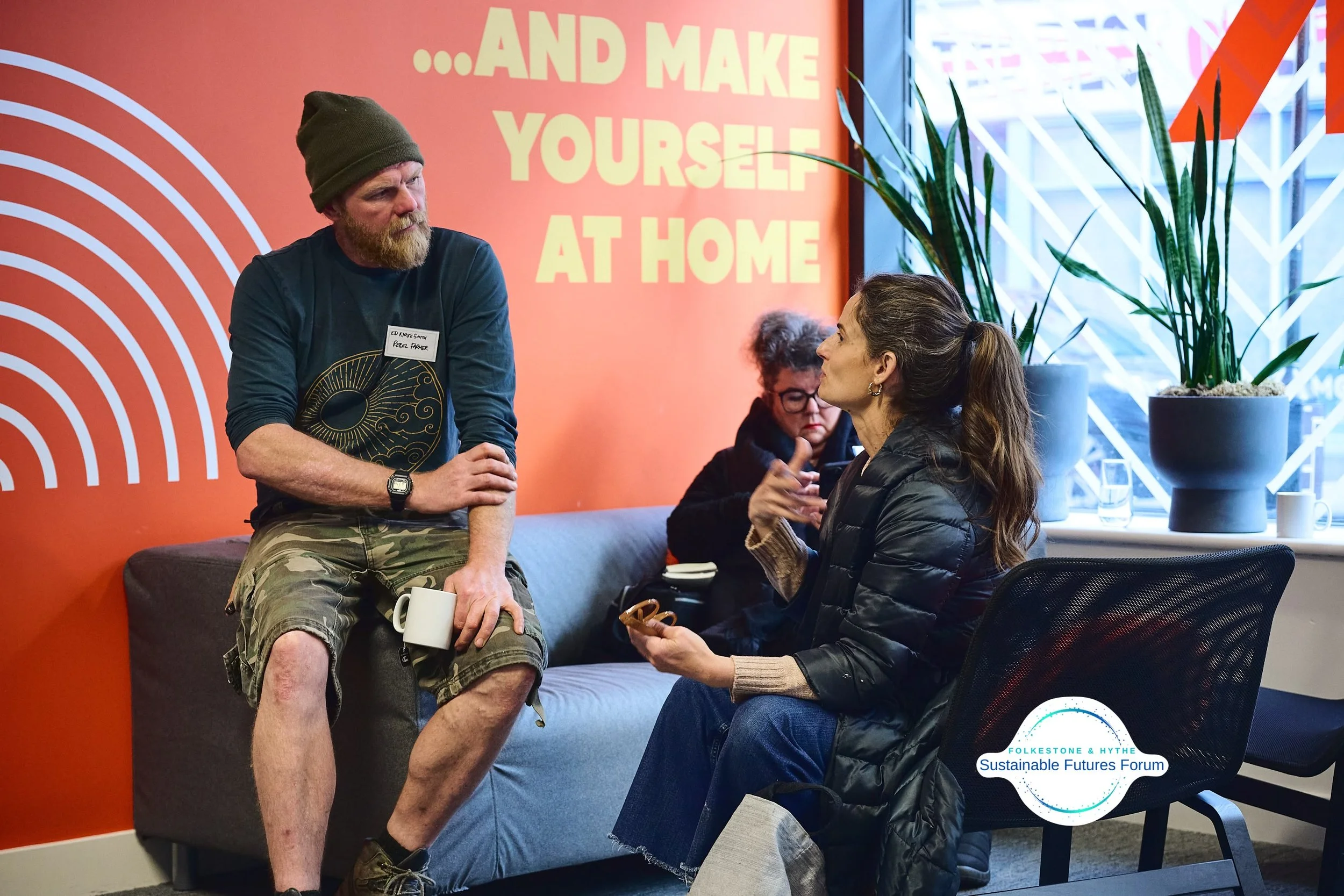 Two women and a man having a conversation inside a modern office or conference room. The woman with brown hair tied in a ponytail is sitting on a chair, wearing a black jacket, and talking animatedly. The man, with a beard and beanie, is sitting on a