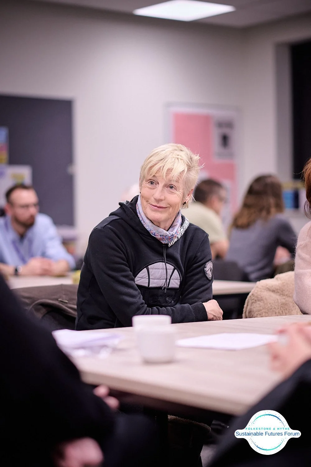 A woman with short blonde hair wearing a black hoodie with graphic designs, sitting at a table in a classroom or conference room, engaged in a discussion. Other people are in the background, some blurred, with a logo of Sustainable Futures Forum in t