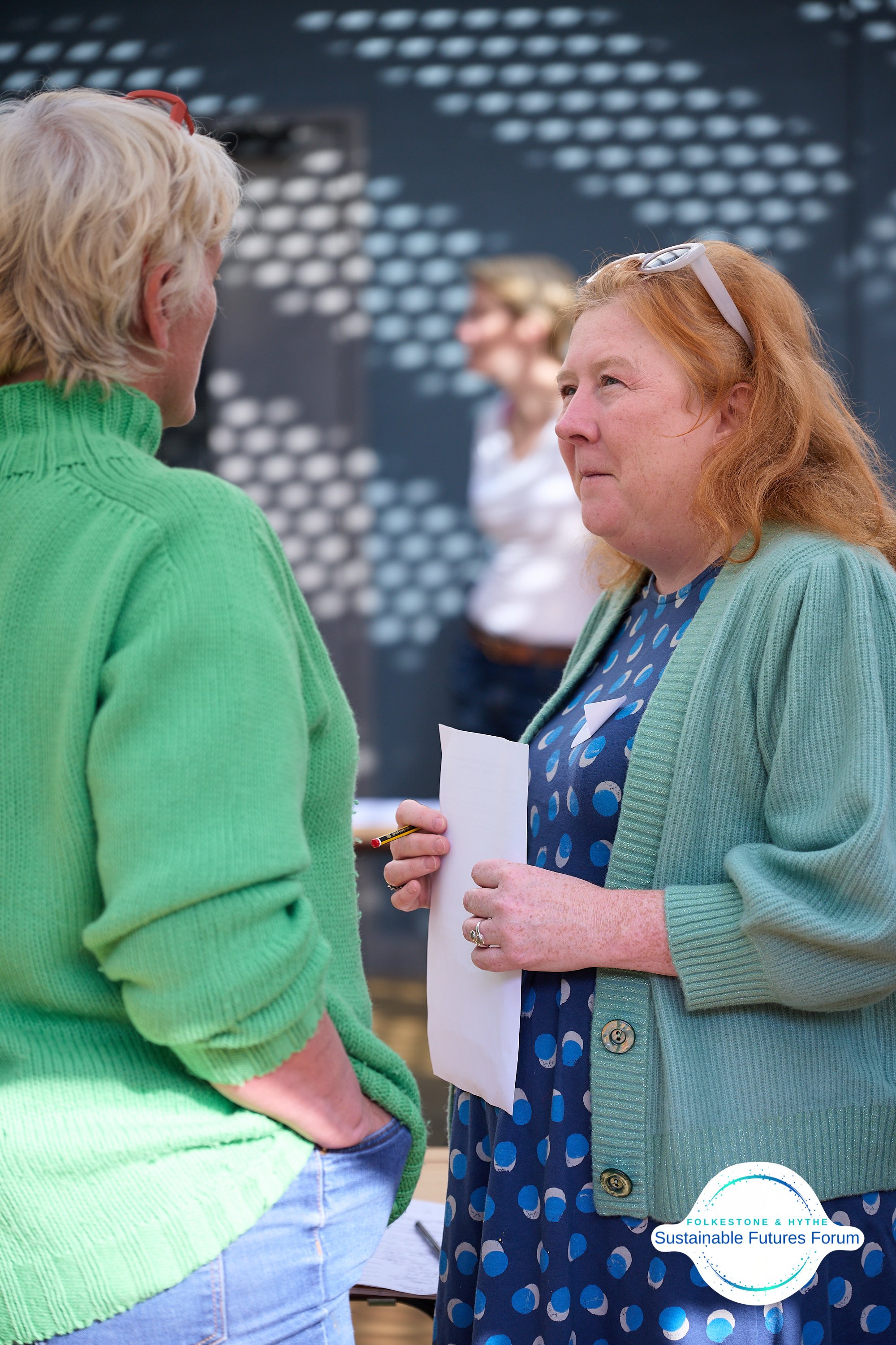 Two women engaged in conversation at the Sustainable Futures Forum, with a blurred background and a logo in the bottom right corner.