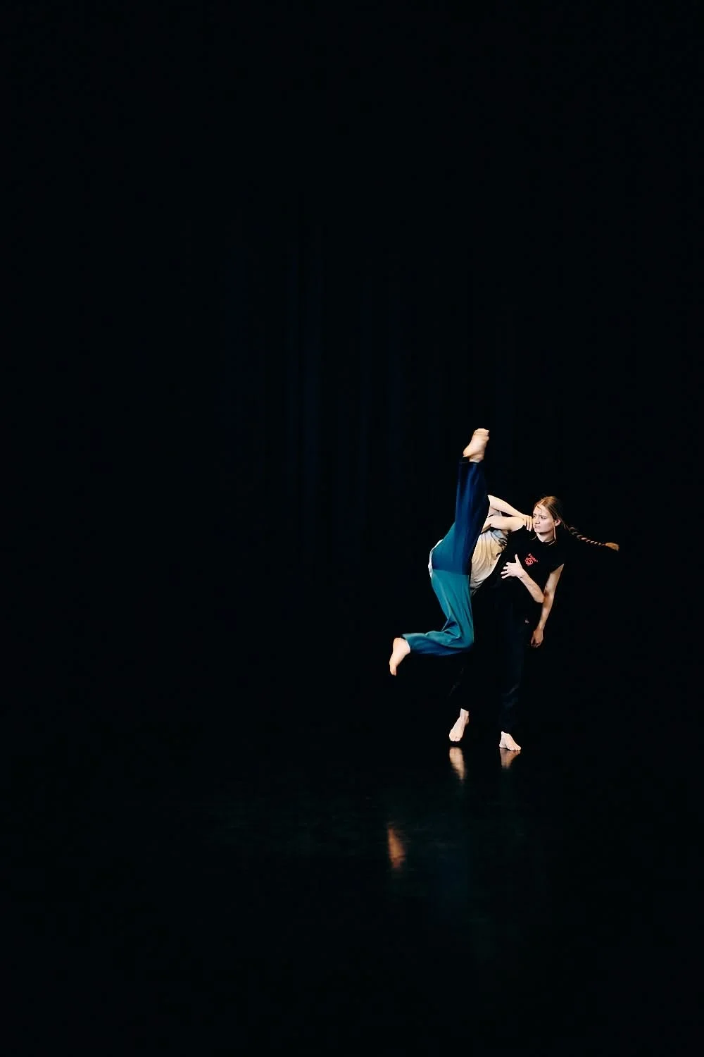 Two dancers performing on a dark stage, one lifting the other mid-air with a black background.