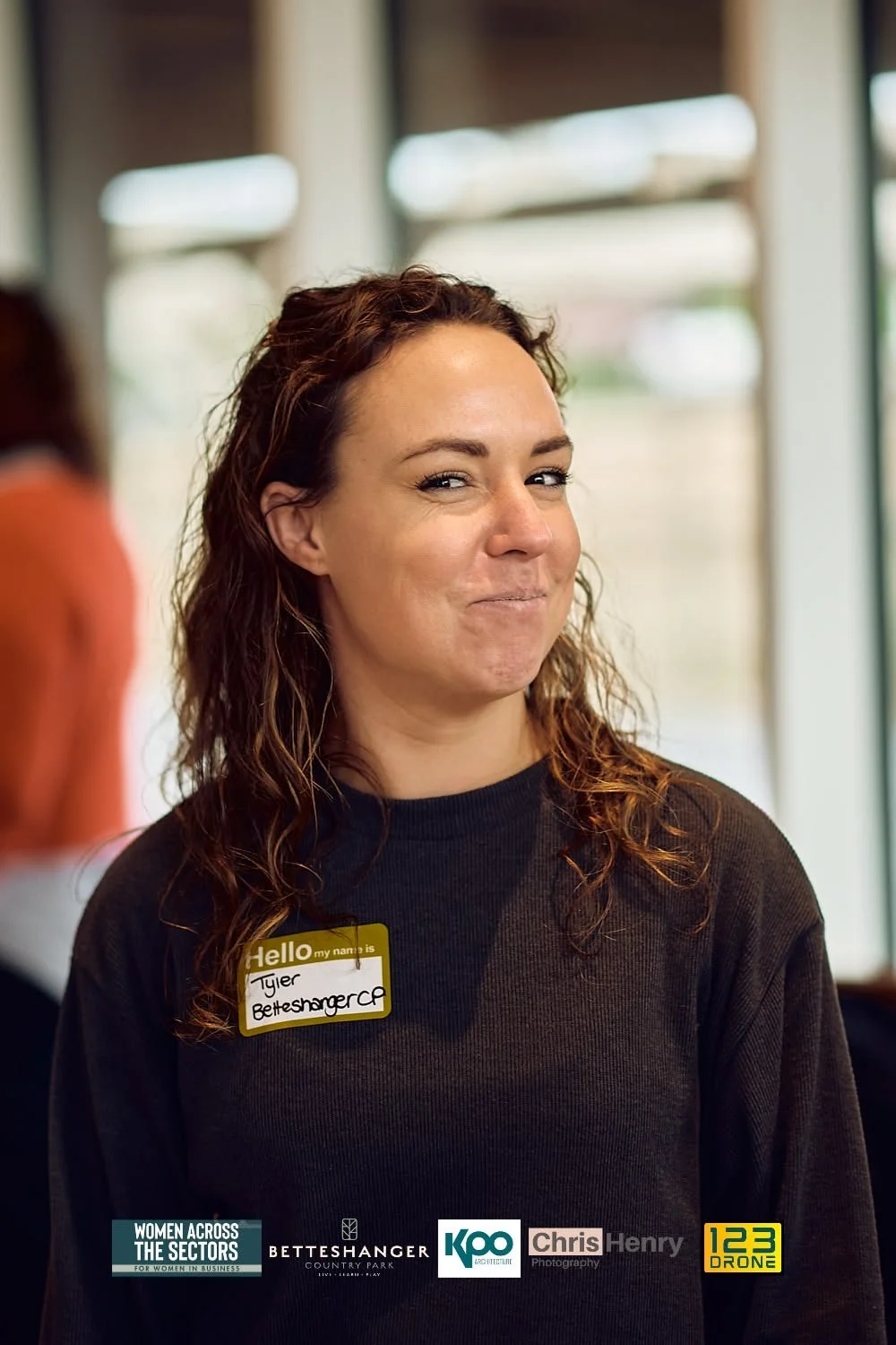 A woman with wavy brown hair smiling slightly, wearing a black shirt with a yellow name tag that says 'Hello, my name is Tyler Betteshanger.' Behind her are large windows, and there are logos at the bottom for 'Women Across the Sectors,' 'Betteshange