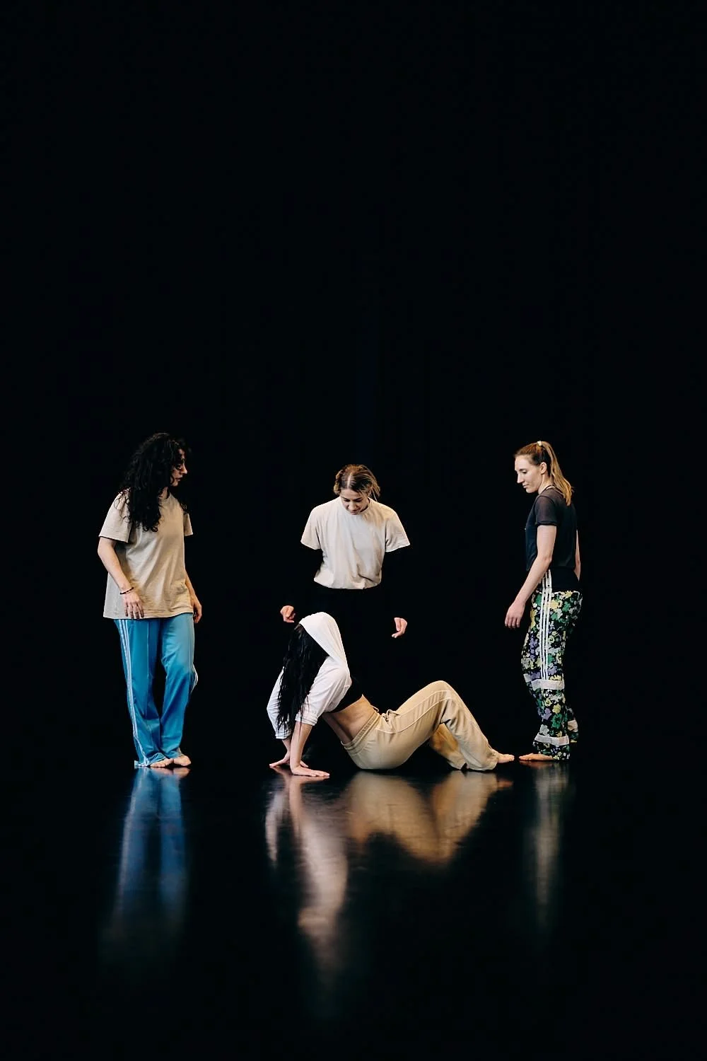 Four women in casual clothing are on a dark stage, with one woman crawling on the floor and the others standing around her, observing.