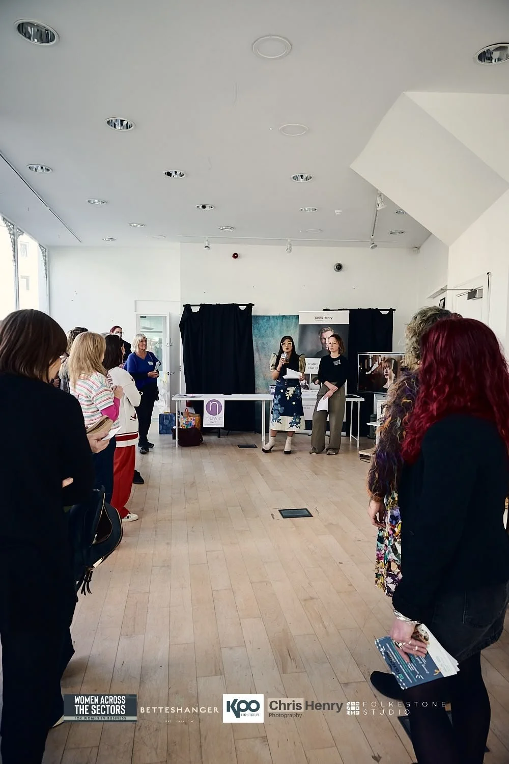 Women attending a presentation or workshop in a bright room with wooden floors, black curtains, and display banners in the background.
