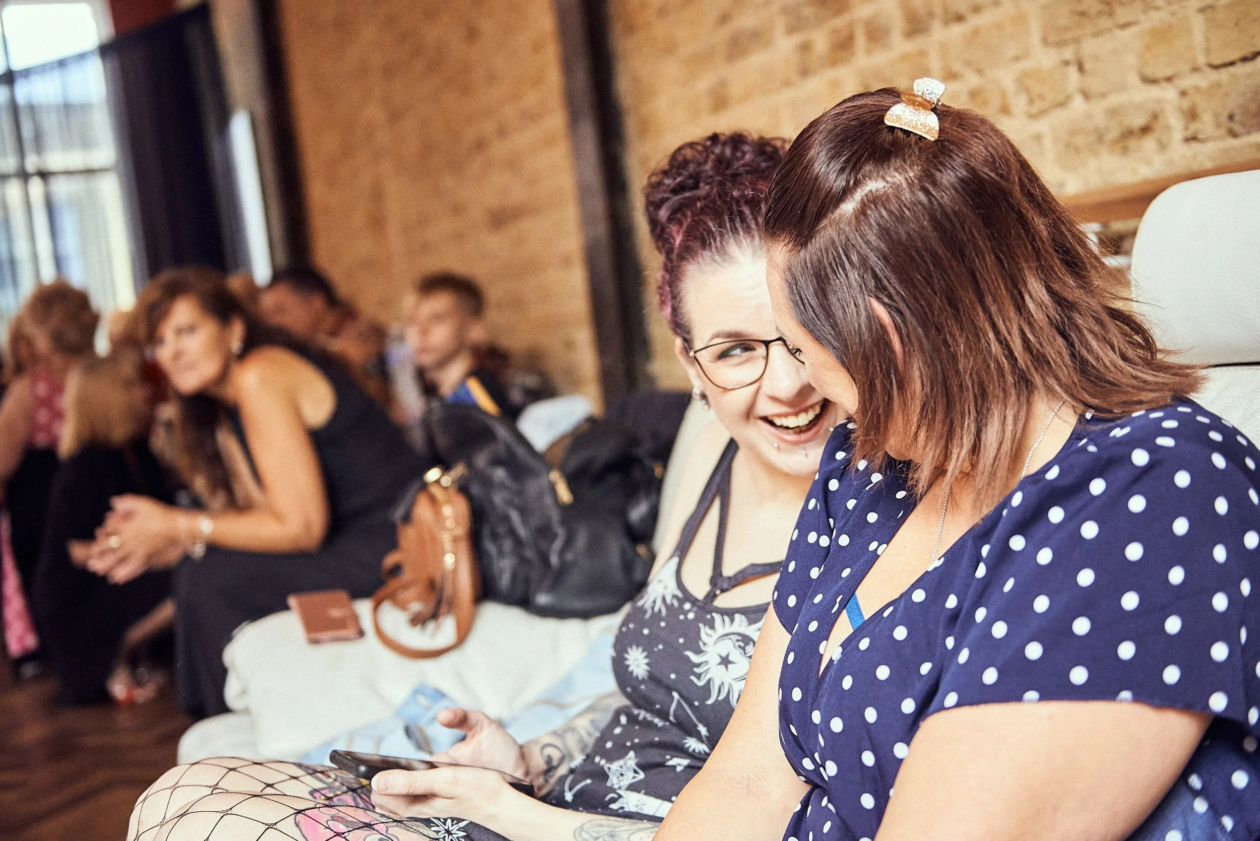A group of women sitting against a brick wall, engaged in conversation and smiling, with some using their phones.
