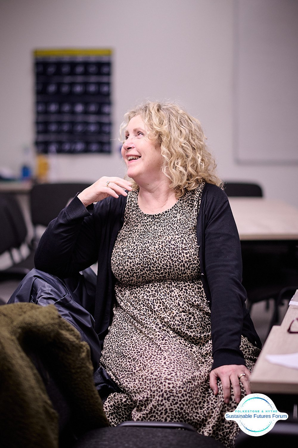 A woman with curly blonde hair, wearing a leopard print dress and black cardigan, smiling and sitting in a classroom or conference room.
