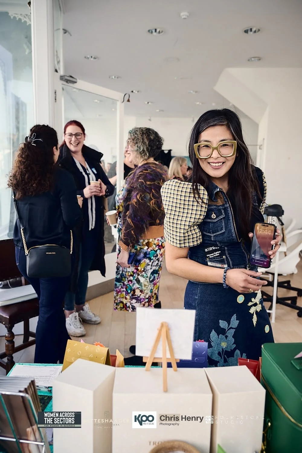 A smiling woman with glasses holding a phone at a social event, with a group of women talking in the background.