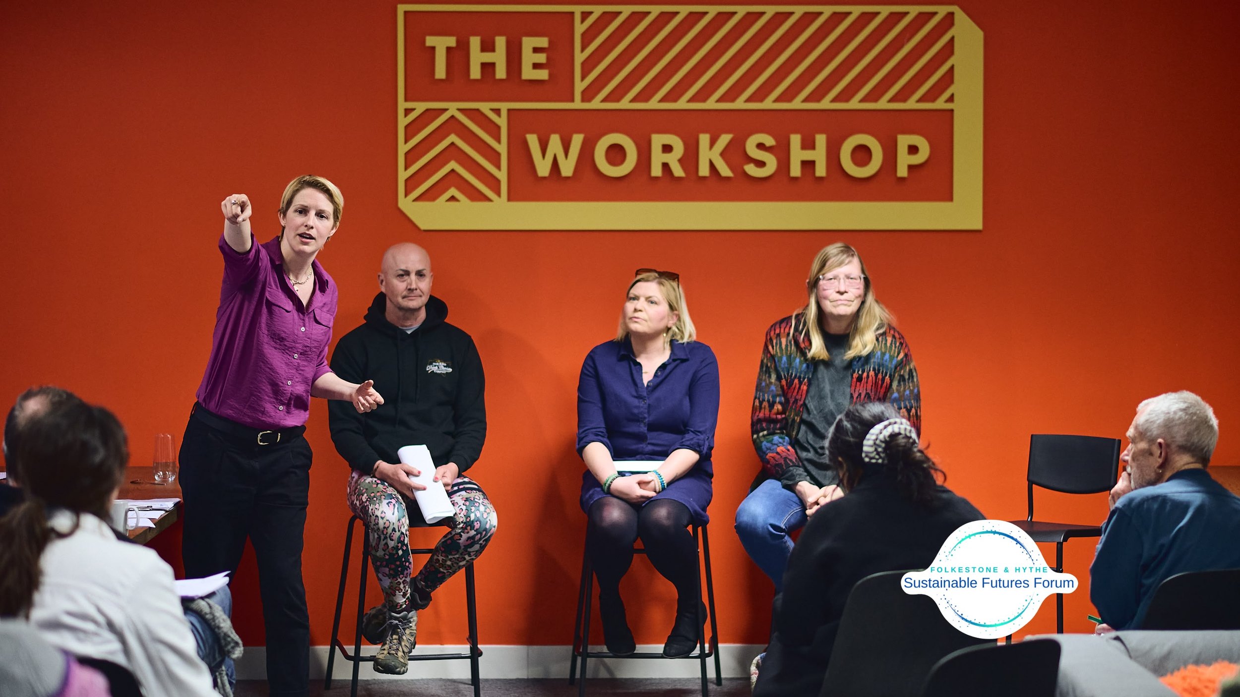 A woman in a purple shirt stands speaking to an audience at The Workshop event, with four people seated on chairs against an orange wall and a logo for the Folkestone & Hythe Sustainable Futures Forum.