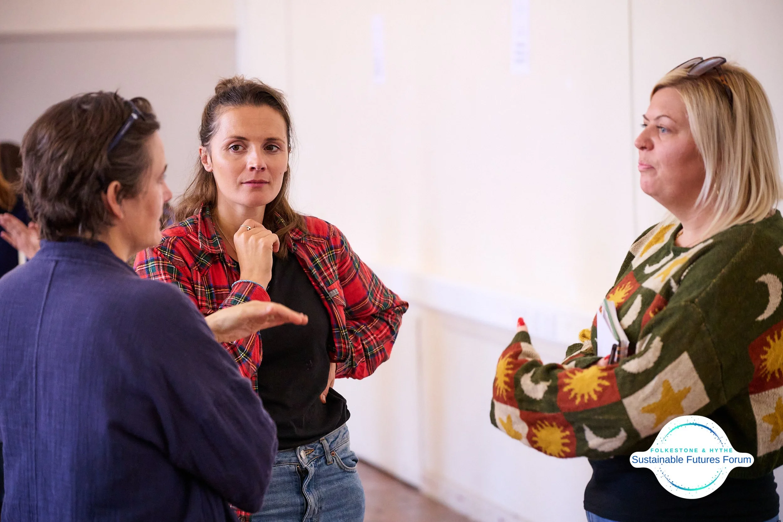 Three women engaged in conversation at an indoor event, with a white wall and a logo reading 'FolkesTone & Hythe Sustainable Futures Forum' in the corner.