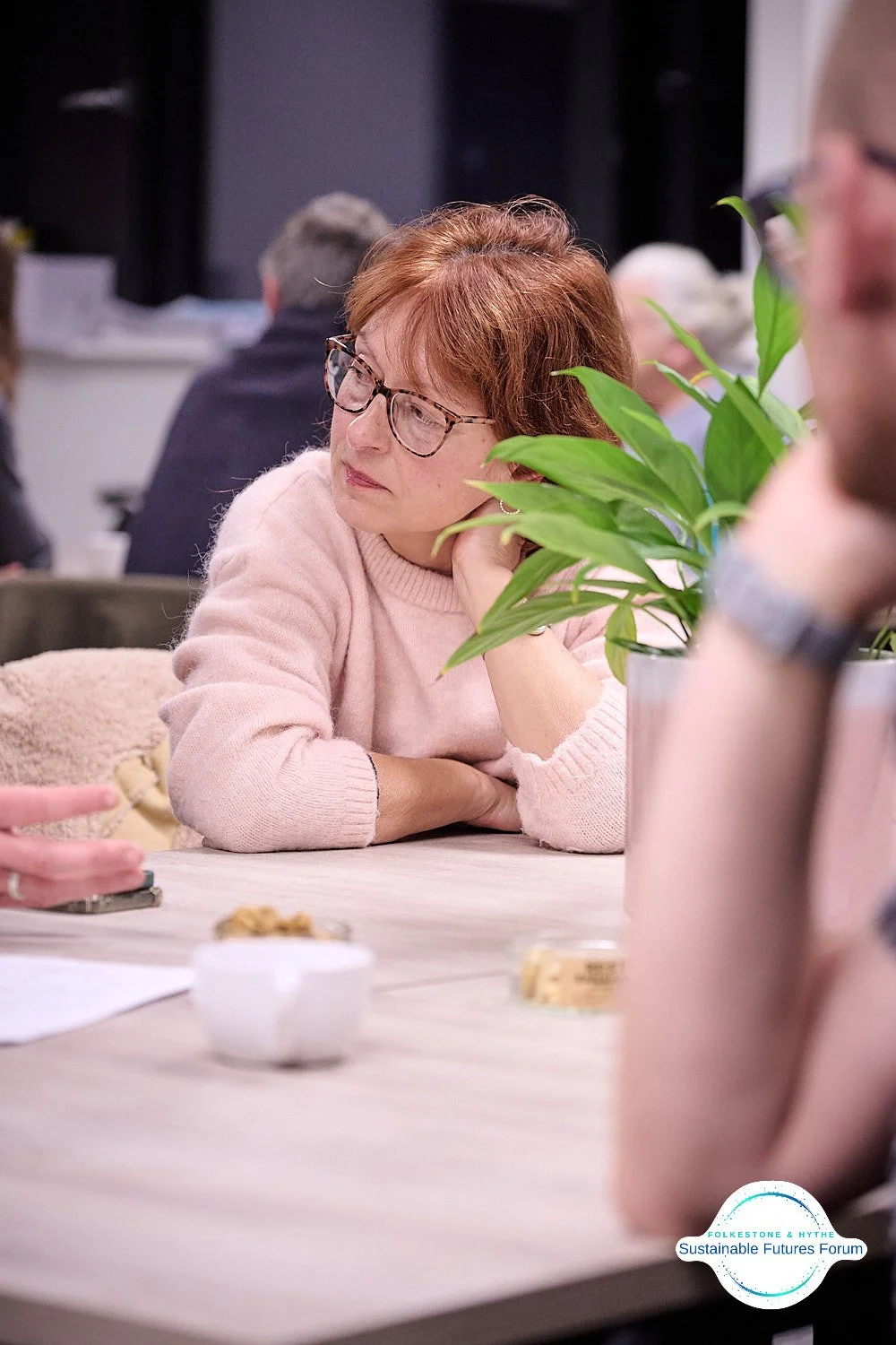 A woman with glasses and reddish-brown hair wearing a pink sweater is sitting at a table, resting her chin on her hand, and listening attentively. There is a potted plant with green leaves on the table, as well as some papers and small containers. In