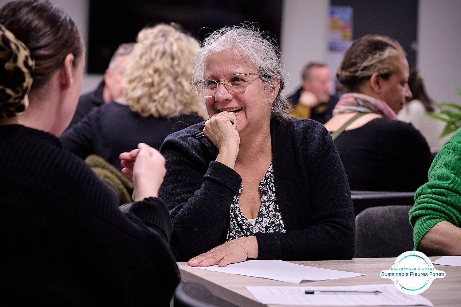 A woman with gray hair, glasses, and a black cardigan smiling and talking with another woman during a forum meeting.