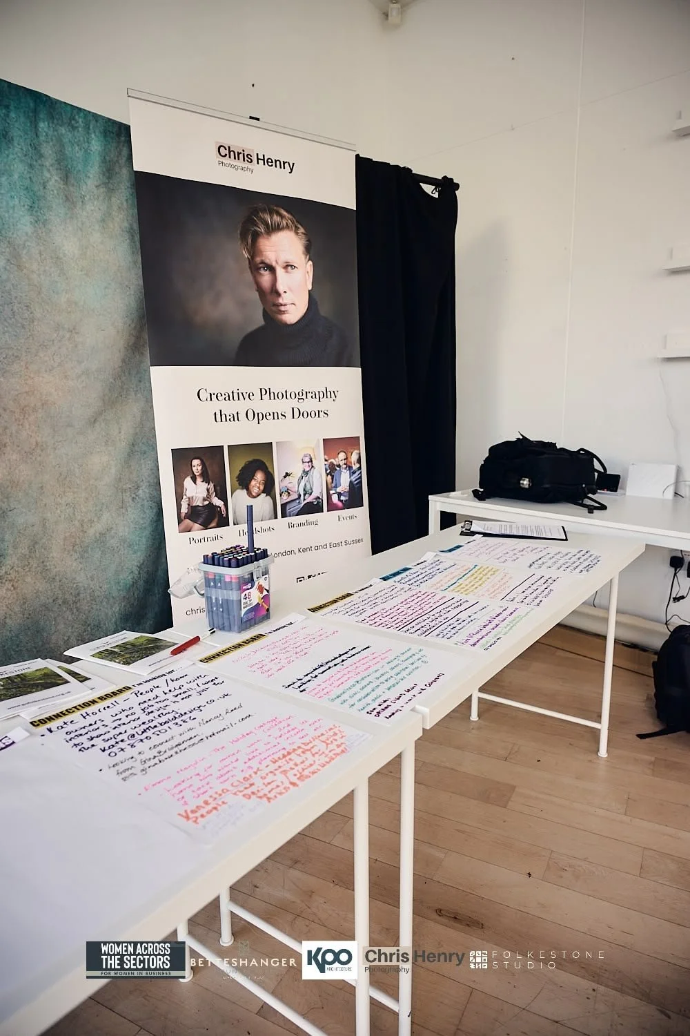 Desk with colorful handwritten notes, open books, and a pack of markers, next to a large vertical banner advertising Chris Henry Photography, with pictures of diverse people and the slogan 'Creative Photography that Opens Doors'.