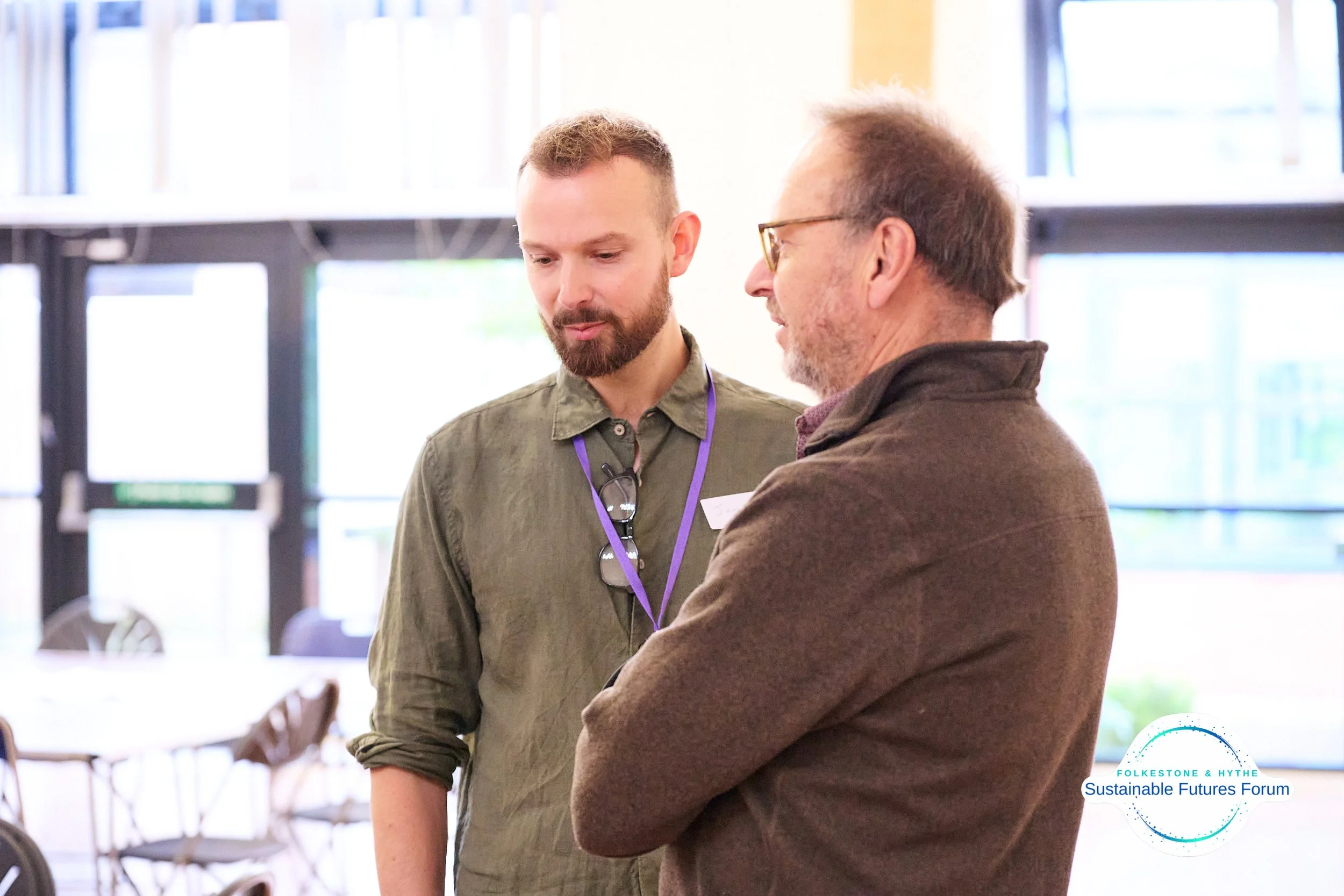 Two men engaged in conversation at the Sustainable Futures Forum event. One is young with a beard and wearing a green shirt, the other older with glasses and a brown jacket. Indoor setting with windows in the background.