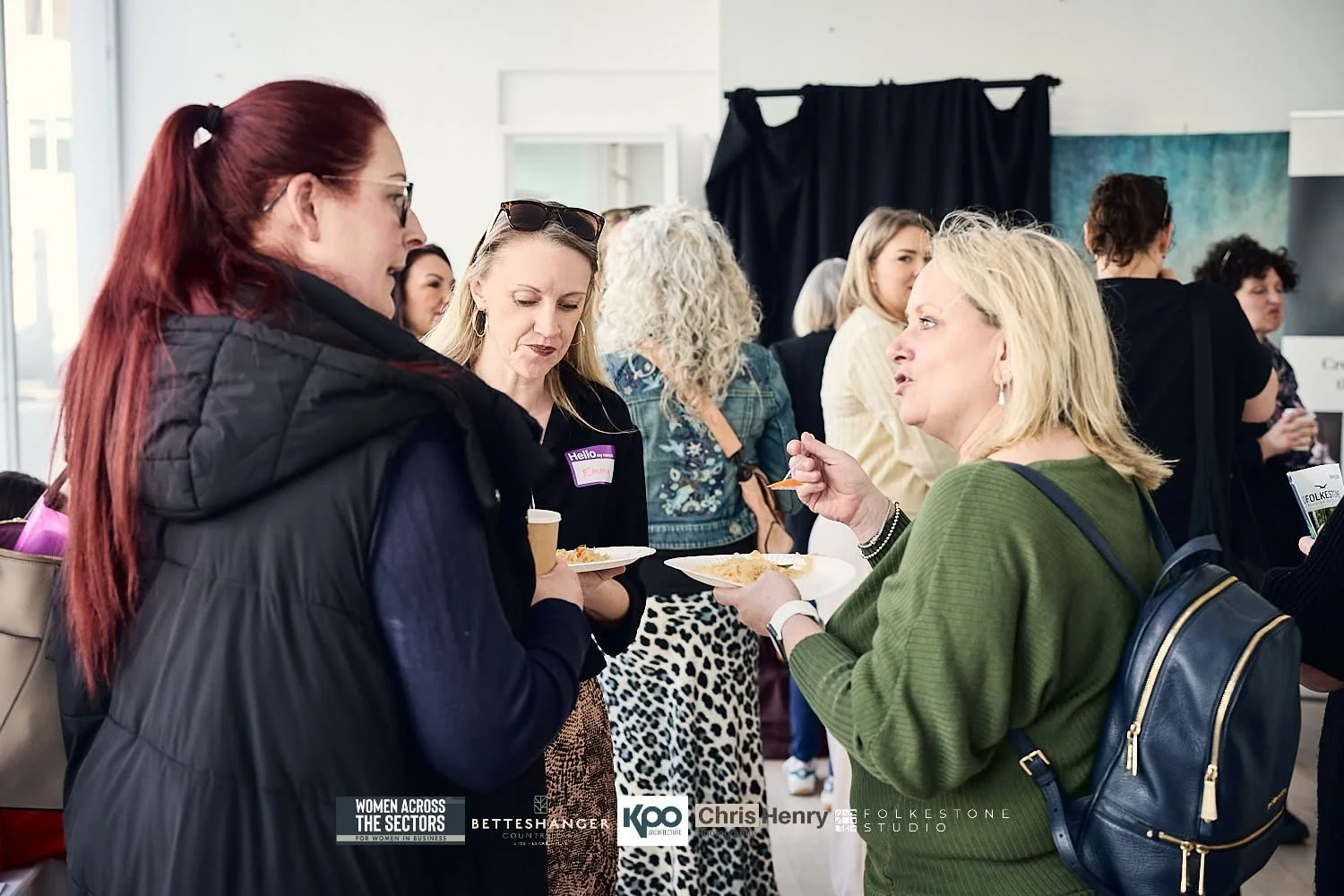Women talking and eating snacks at a social event with many people in the background.