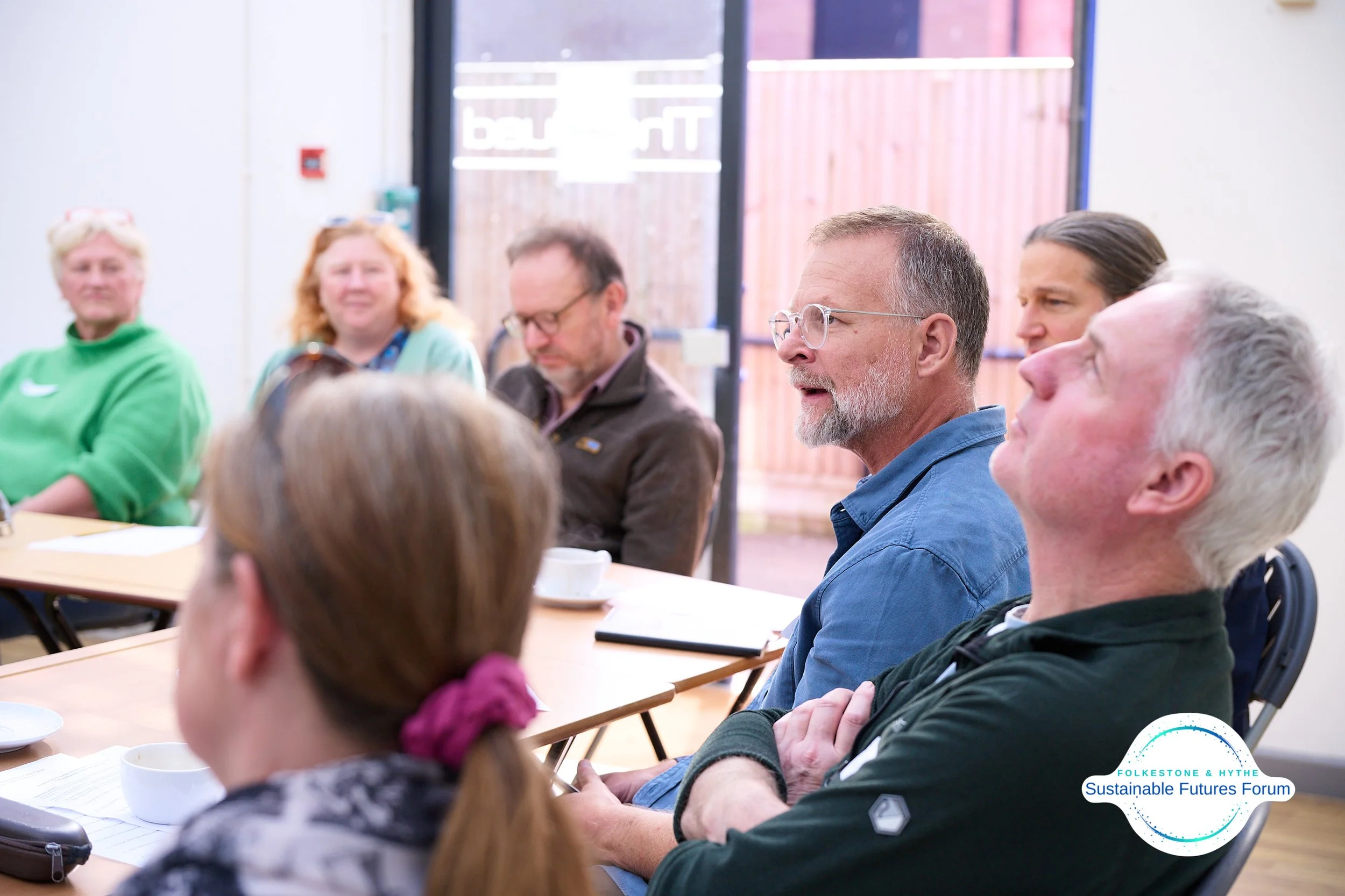 A group of people sitting around a conference table attending a forum or meeting. One man is speaking, and others are listening attentively. There is a sign on the window that reads 'The'.