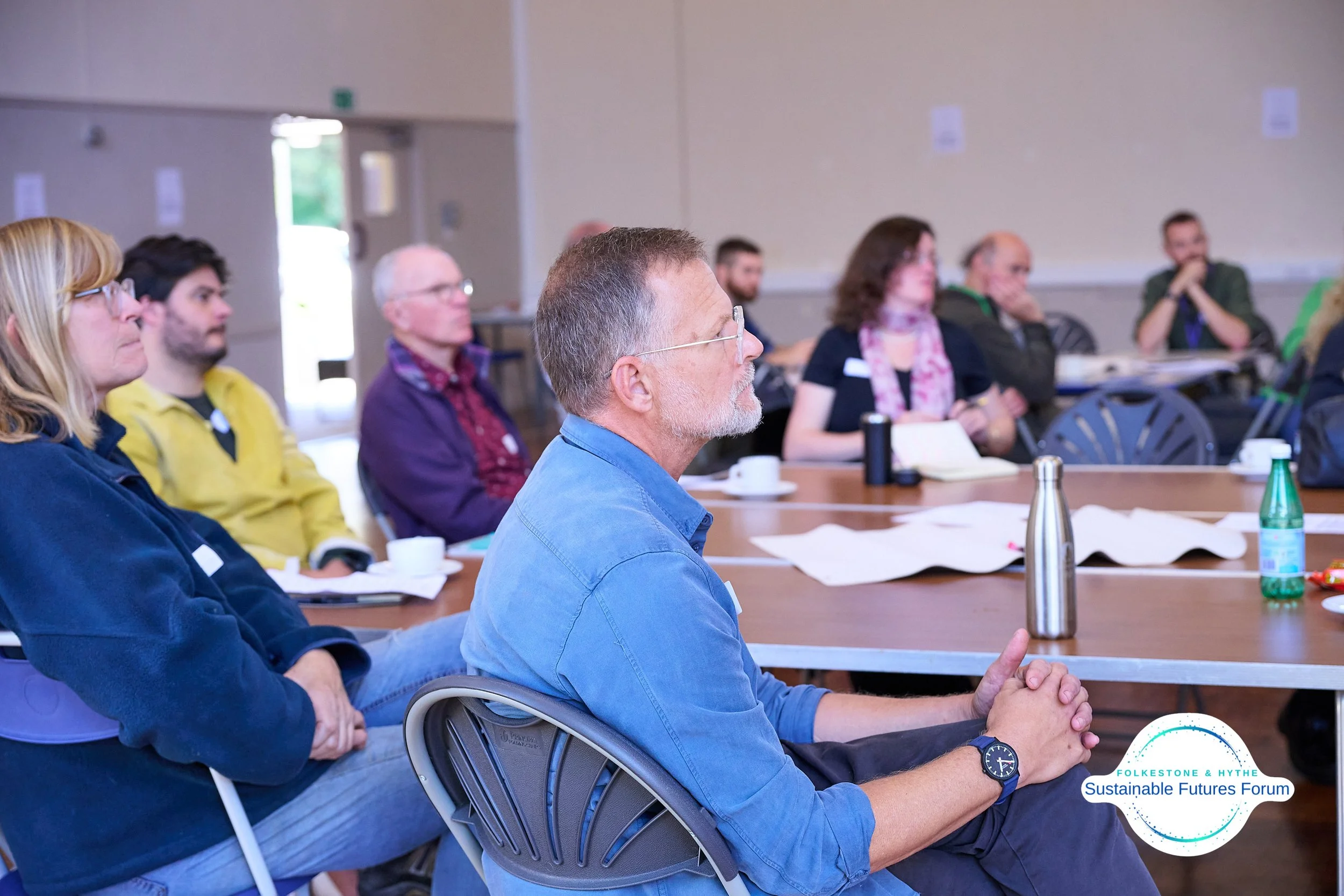 People attending a forum or meeting at a conference table, listening attentively, with notebooks and drinks in front of them, logo in lower right corner reading 'Folketone & Hythe Sustainable Futures Forum'.