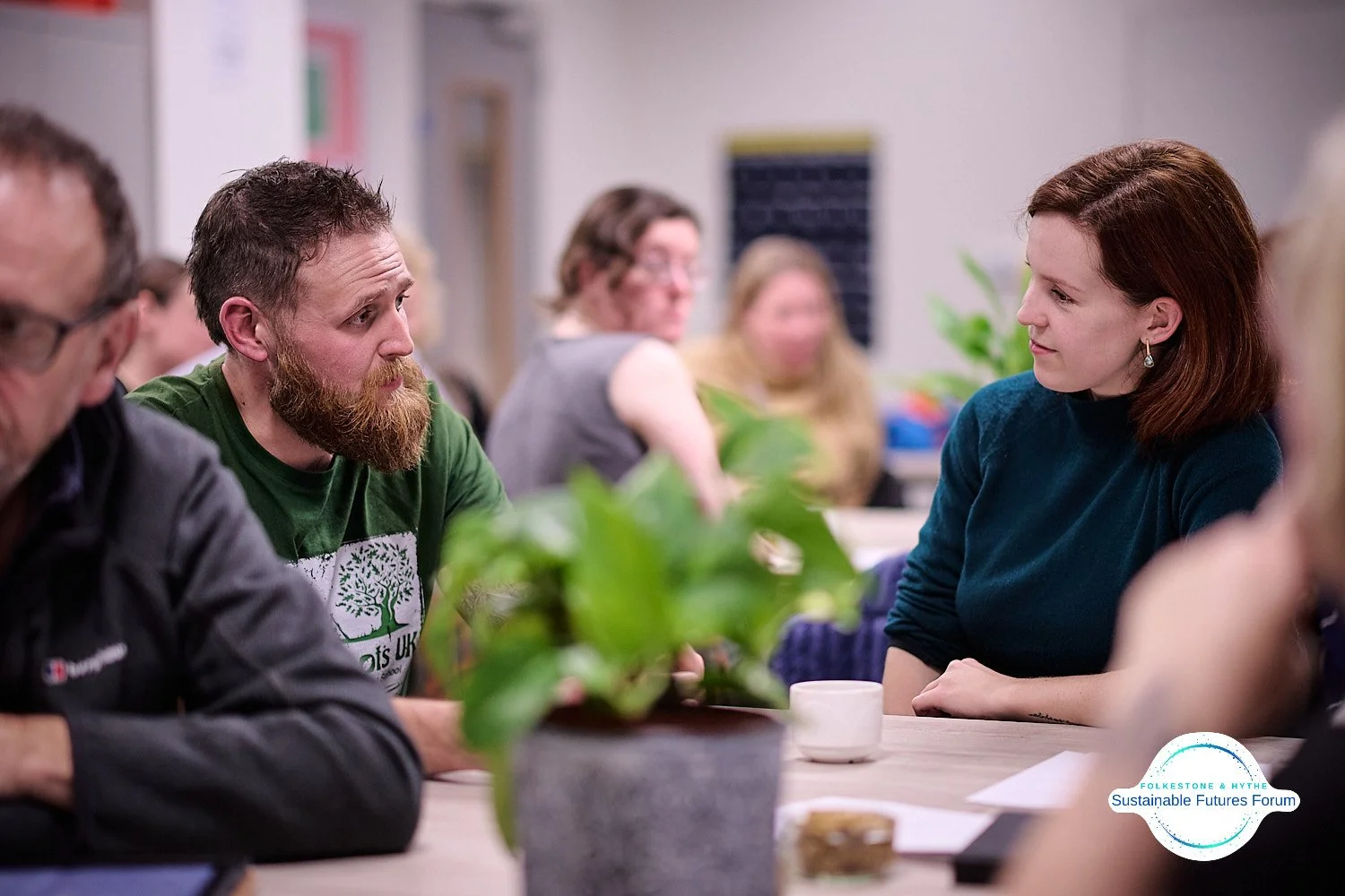 People sitting at a table engaged in conversation, with a woman in a blue sweater and a man with a beard and green shirt talking to each other, in a room with others in the background.