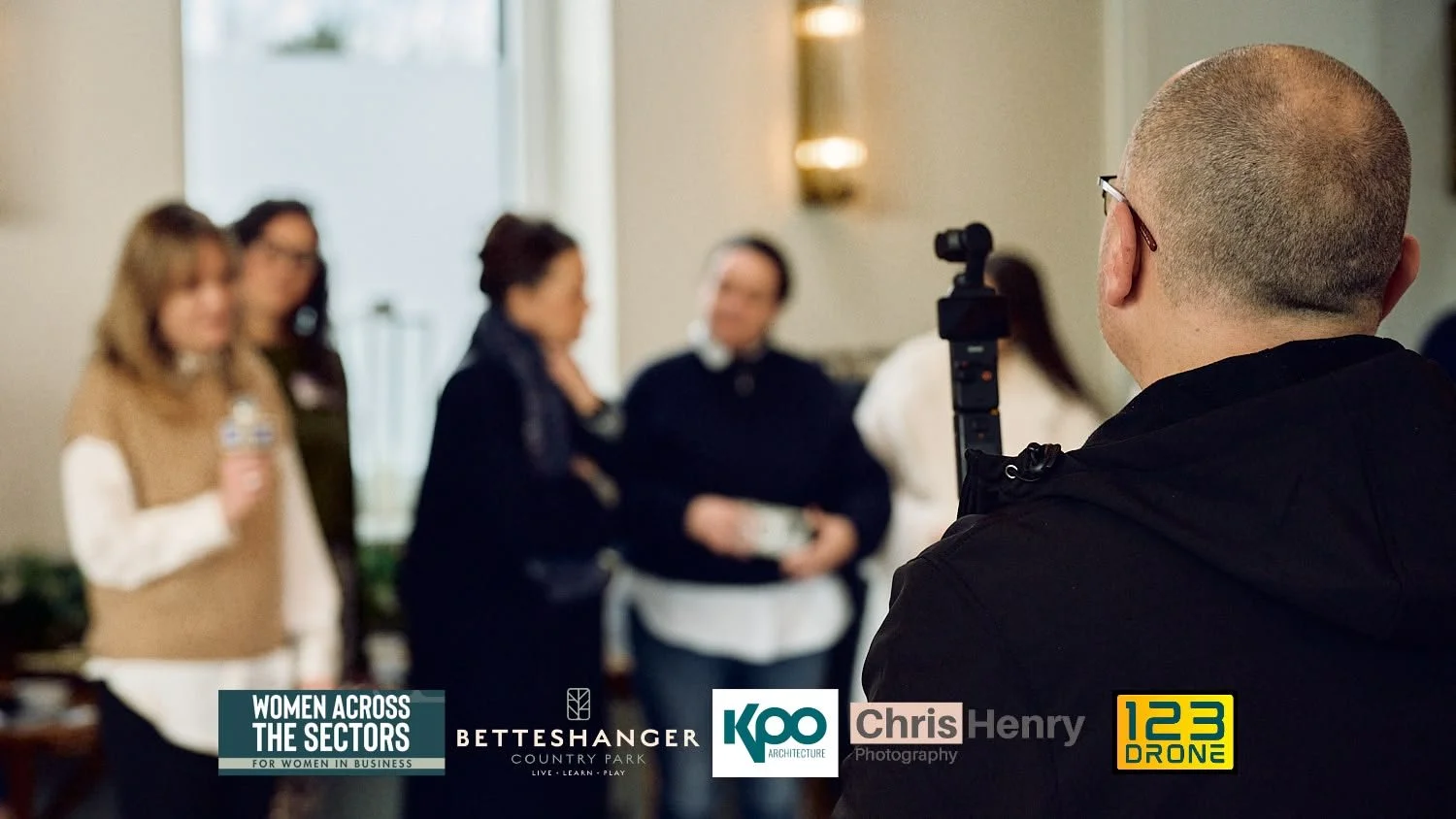 Photographer taking picture of women at Women Across the Sectors event with logos for BETTESHANGER, KPO Architecture, Chris Henry Photography, and 123 Drone visible.