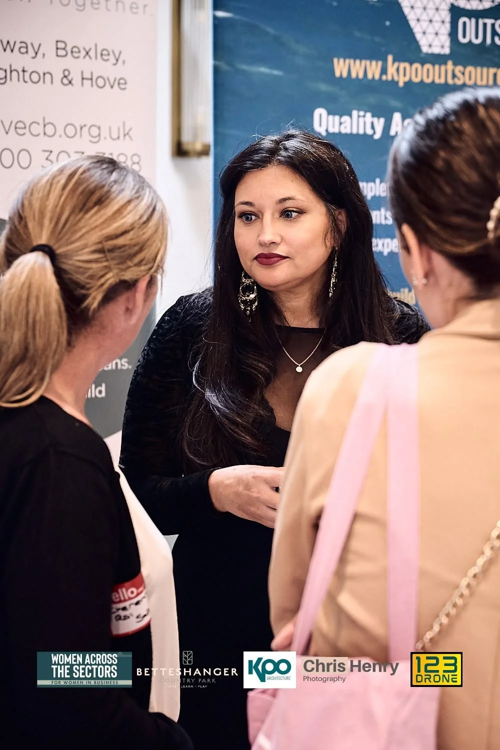 Three women engaged in conversation at a professional event, with banners and logos in the background.