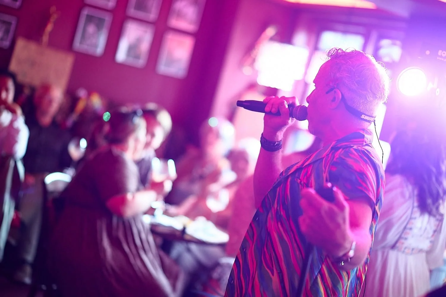 A singer with curly gray hair and glasses performs into a microphone at a lively, dimly-lit venue, with audience members sitting and standing around, under colorful purple and pink lighting.