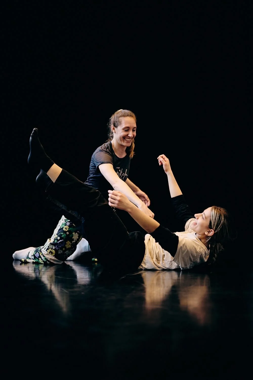 Two women practicing dance or martial arts on a black stage, one woman is lying down and holding the other's hands, both smiling.