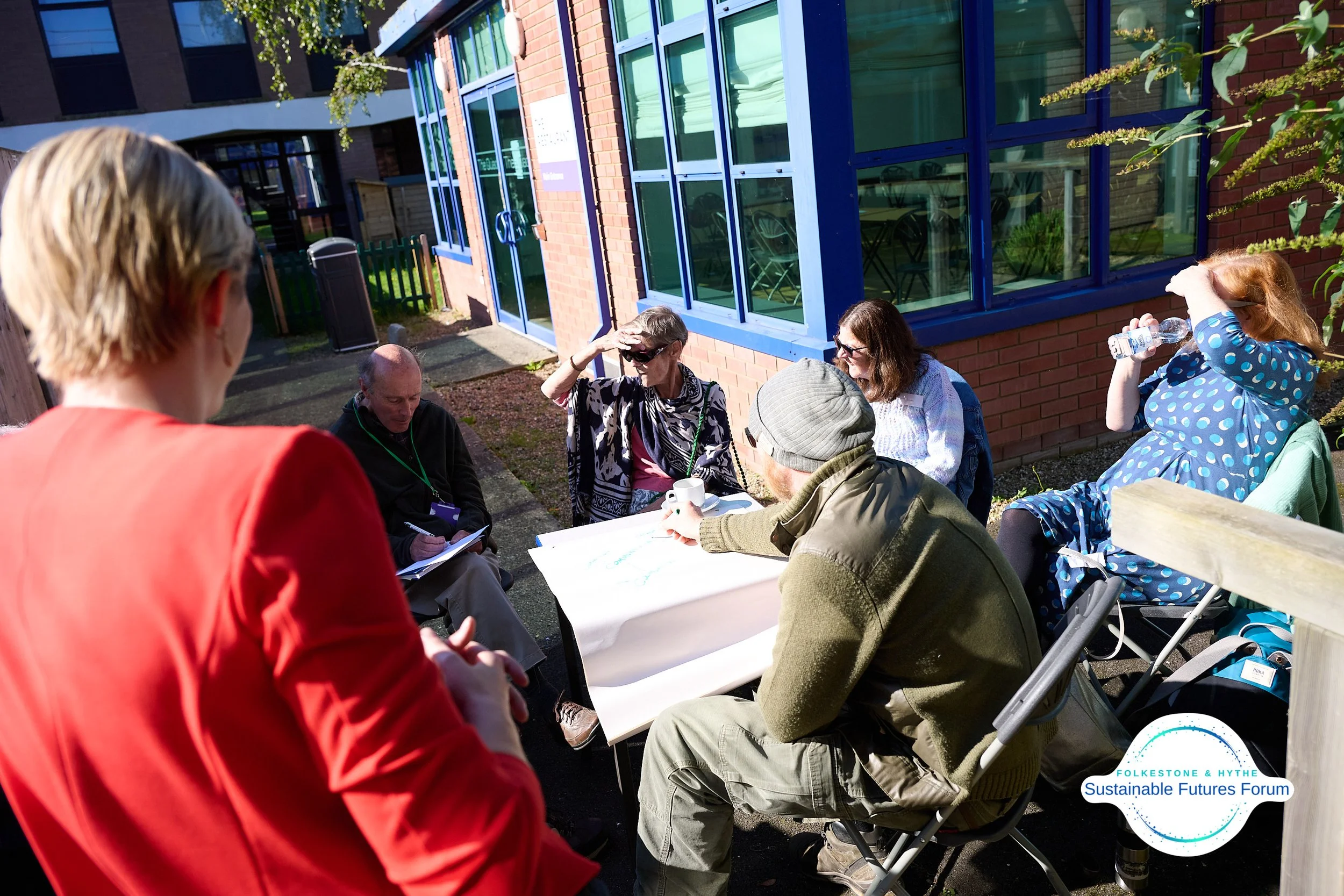 A group of people participating in an outdoor discussion or meeting, seated around a table near a brick building with large blue windows, on a sunny day.