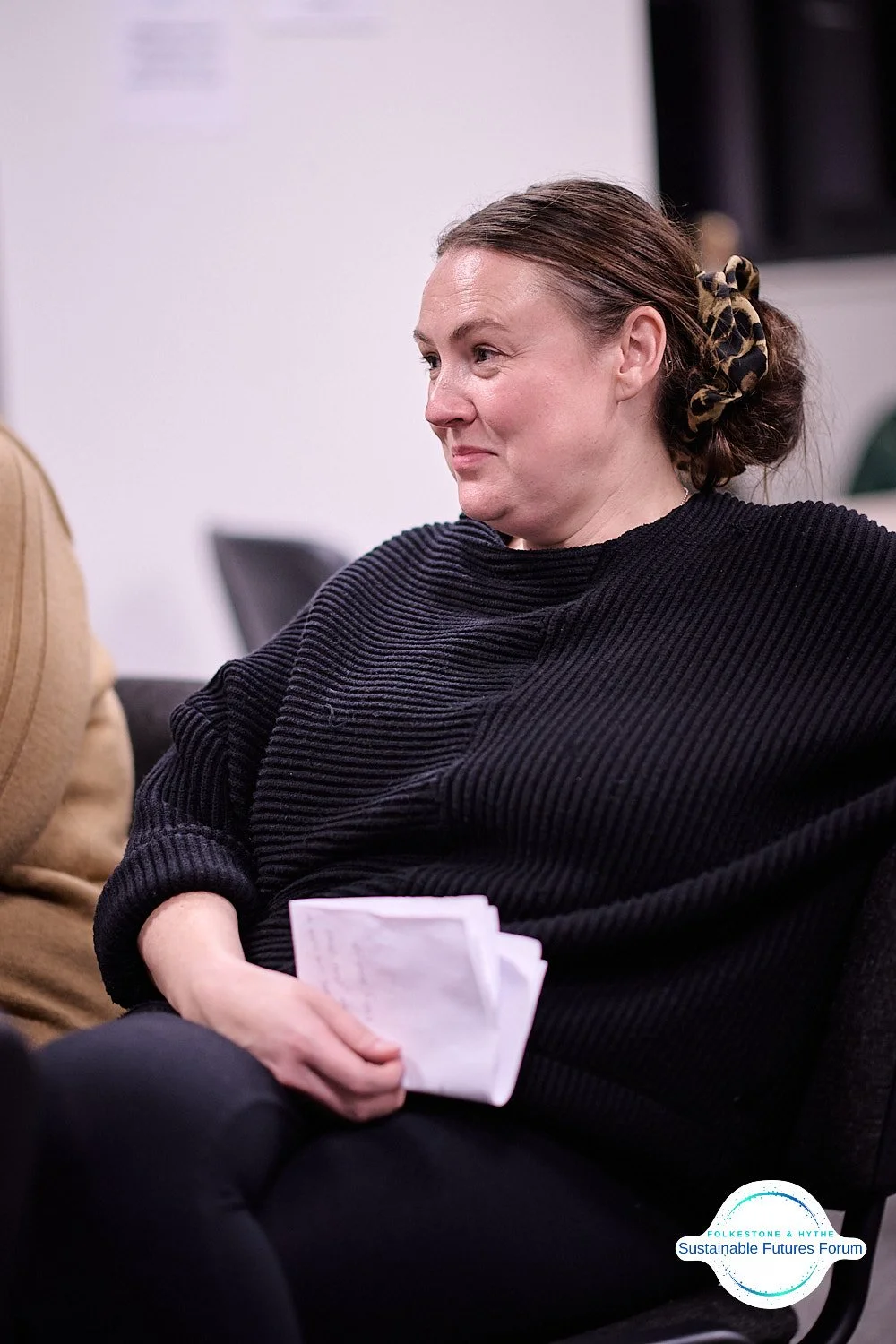 A woman with brown hair tied back with a leopard print scrunchie, wearing a black sweater, sitting on a black couch, holding a notepad or papers, and smiling slightly during a discussion at an indoor event.