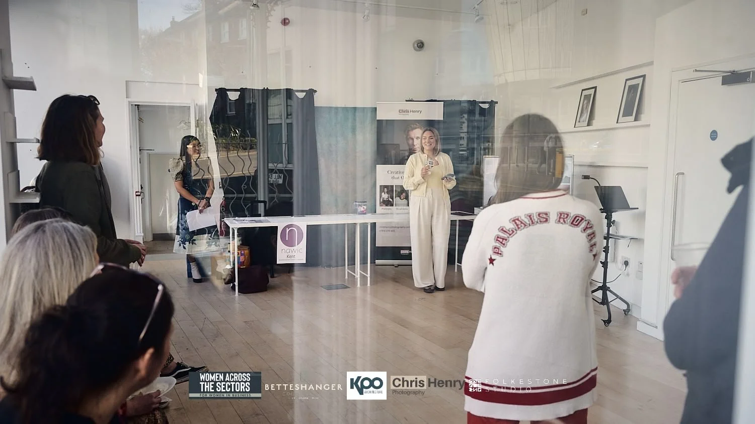 A woman is speaking to an audience through a glass window at a women’s business event, with some people seated and standing inside, and various banners and photos on the walls.