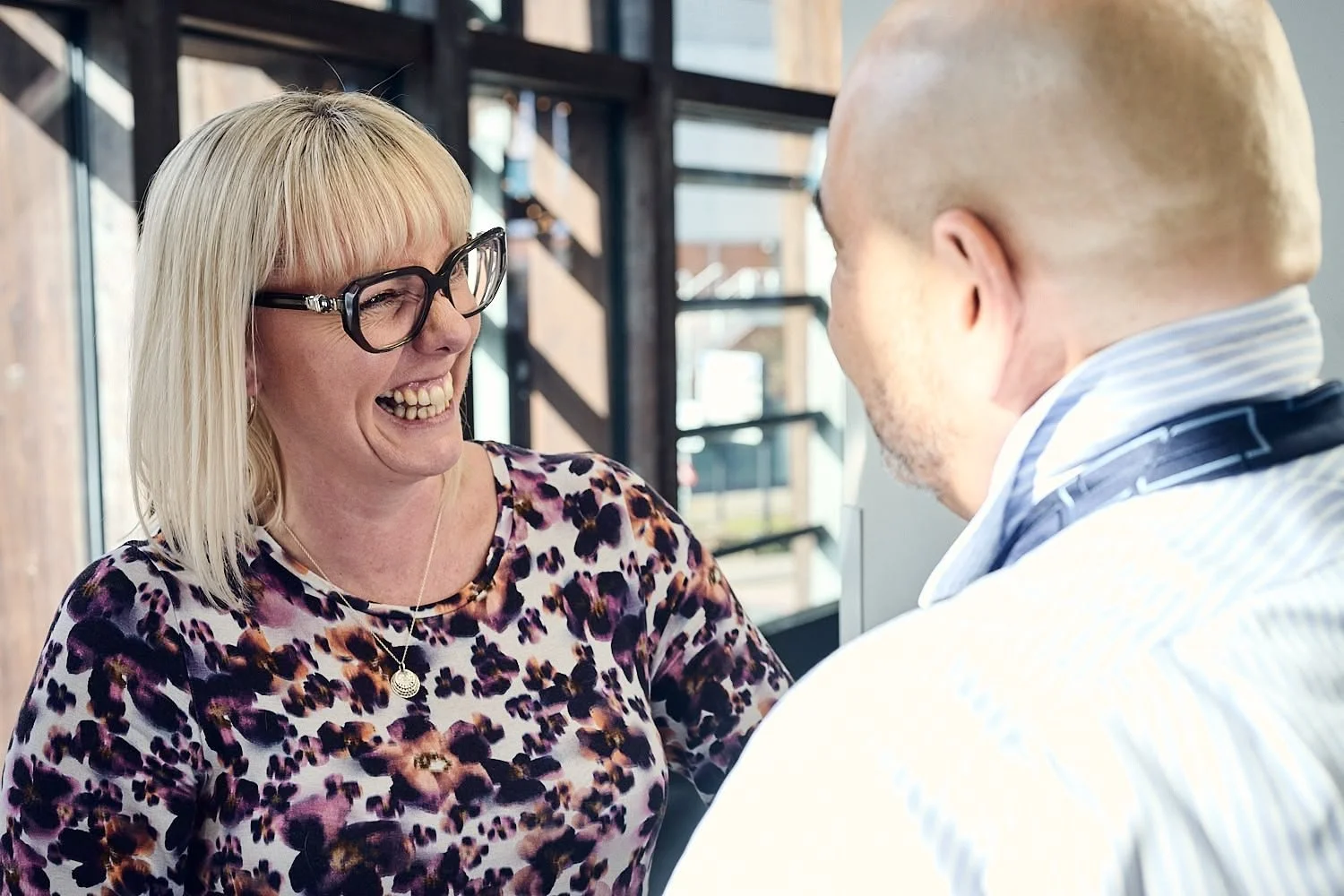 A woman with blonde hair, glasses, and a leopard print shirt smiling and laughing with a person wearing a striped shirt in what appears to be a social or professional setting.