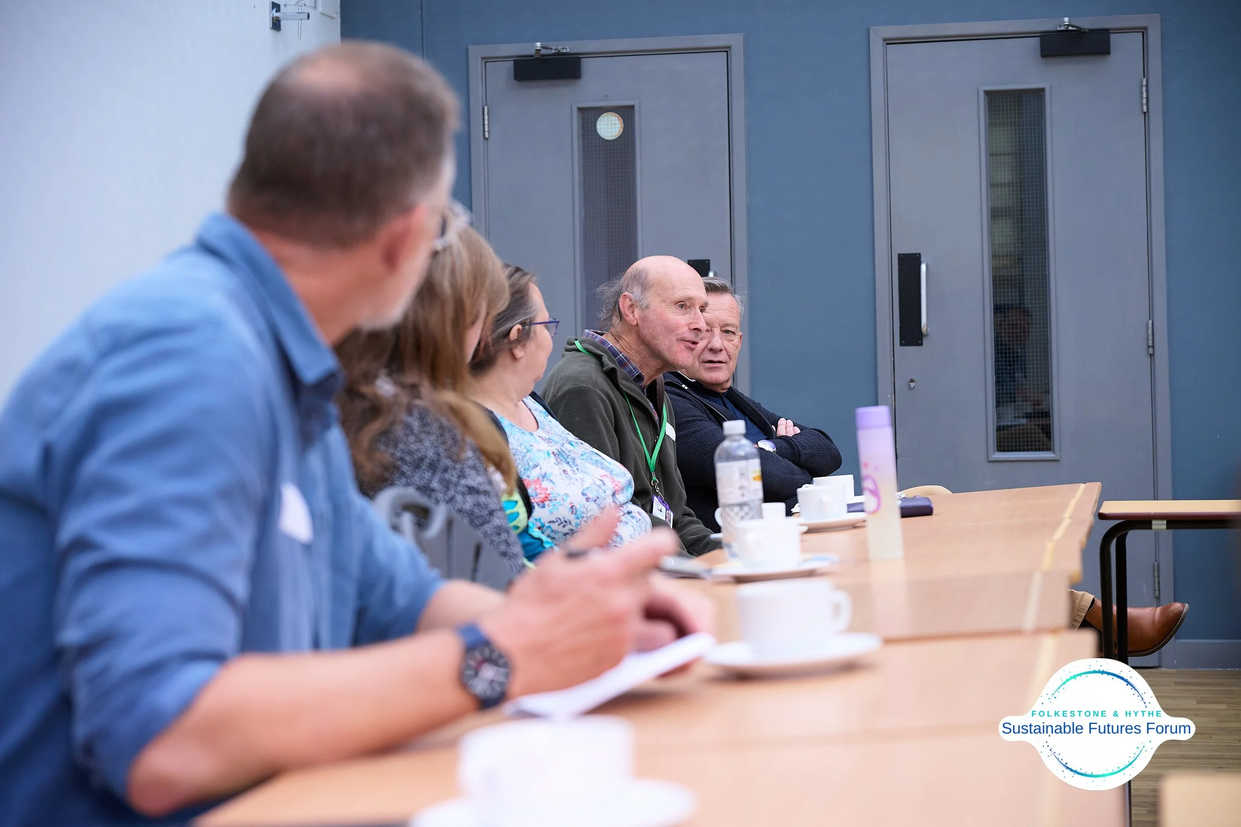 A group of five people sitting at a conference table, engaged in a discussion, with cups and bottles on the table, and a panel discussion happening at the Sustainable Futures Forum.