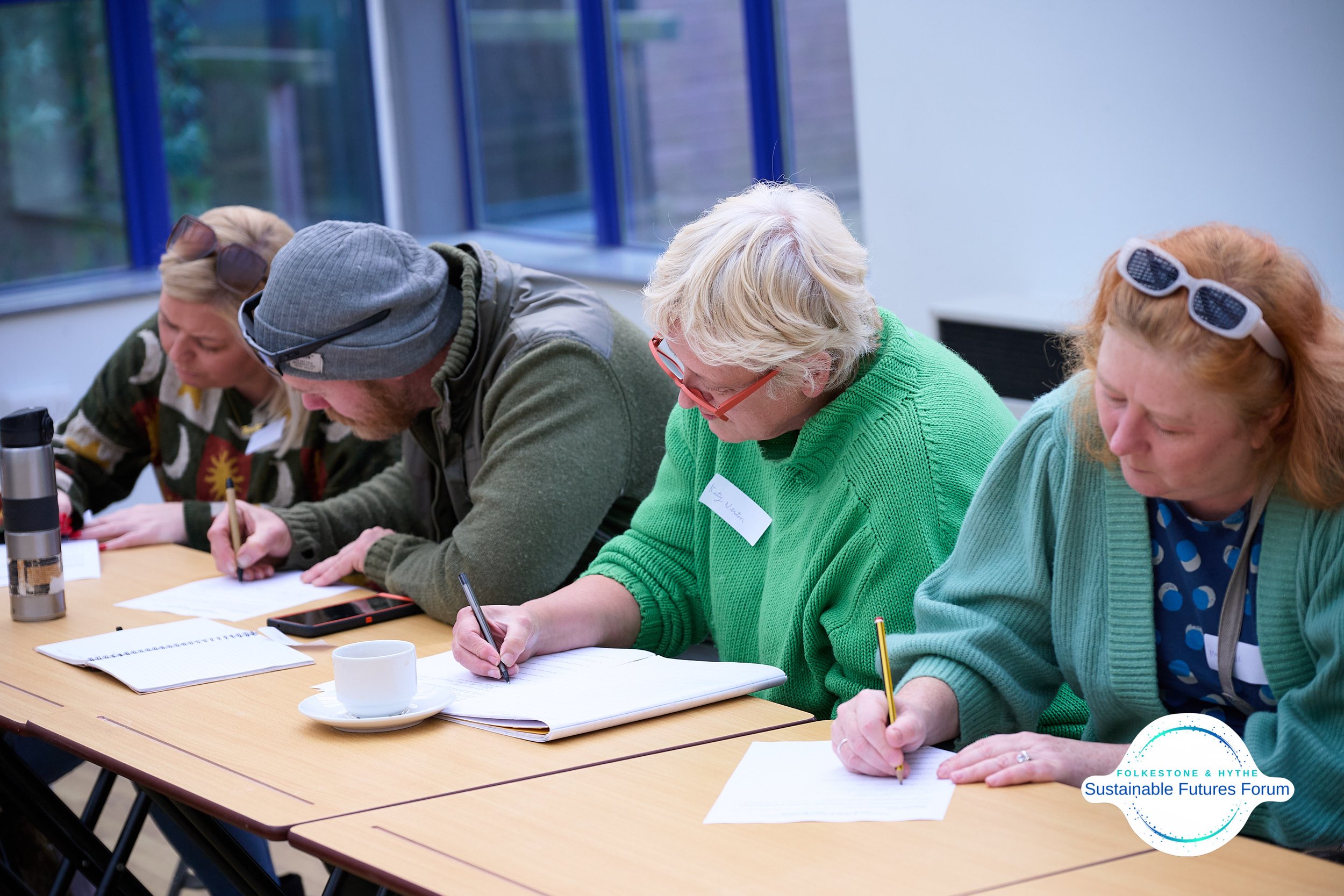 People sitting at a table, writing on paper, during a forum or workshop, with a logo in the bottom right corner indicating 'Folkes Tone & Hythe Sustainable Futures Forum'.