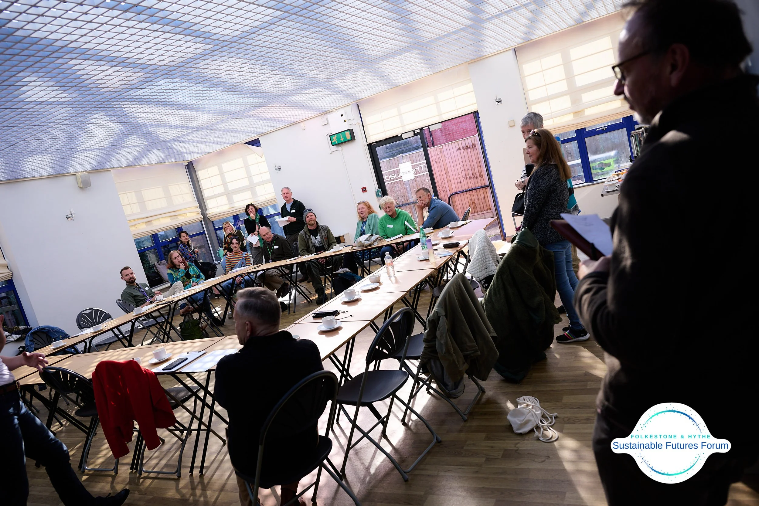 People gathered in a bright conference room with a U-shaped arrangement of tables, listening to a speaker standing in front. Some are taking notes, and there are cups, bottles, and notebooks on the tables. The room has large windows with blinds, and 