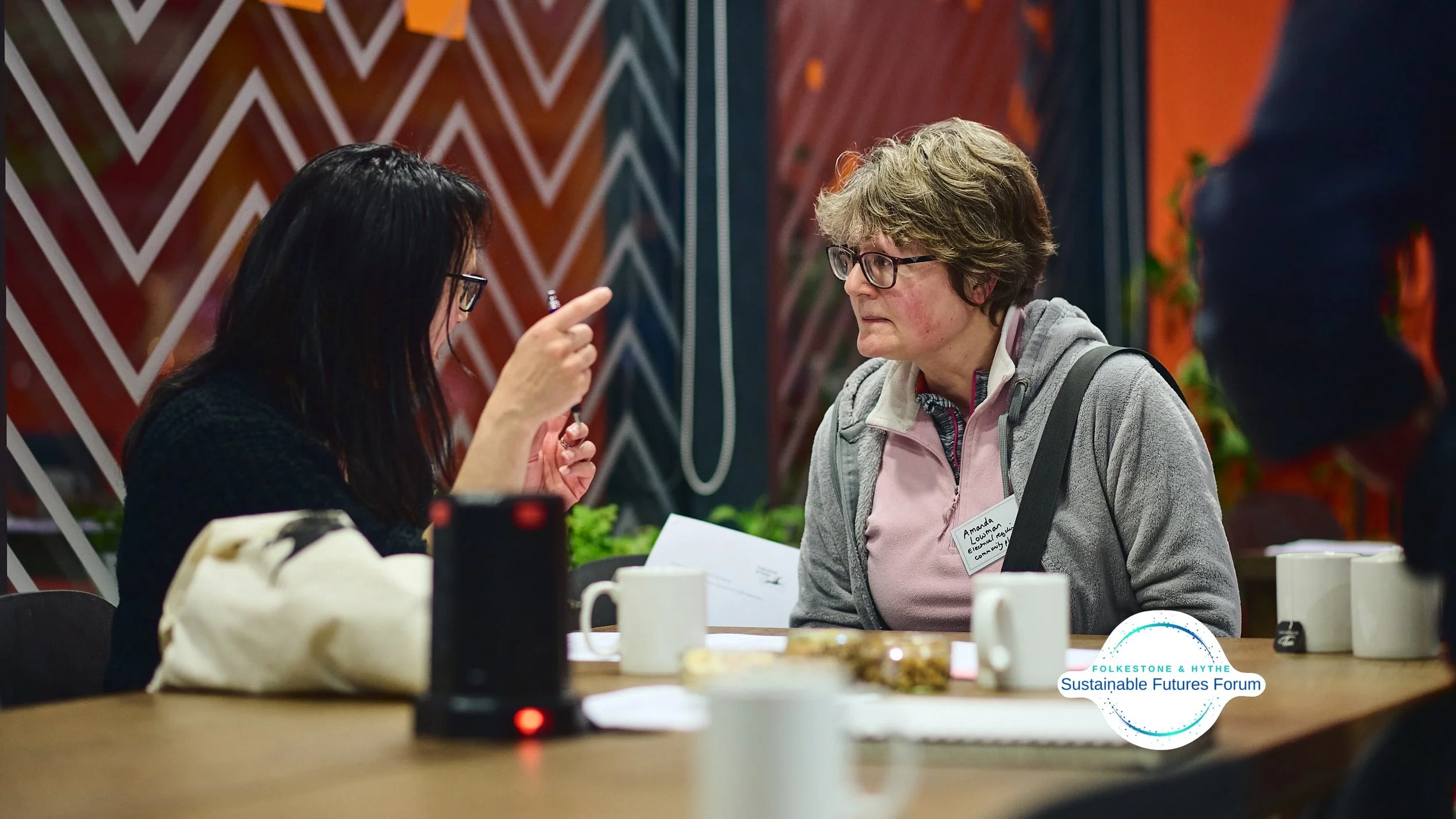 Two women sitting at a table having a conversation in a meeting or discussion setting, with several mugs and papers on the table, and a logo that reads 'Sustainable Futures Forum.'