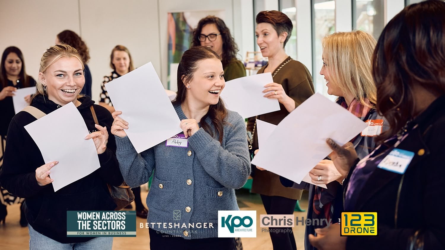 Women engaging in a networking event, smiling and holding papers in a bright, modern indoor space.