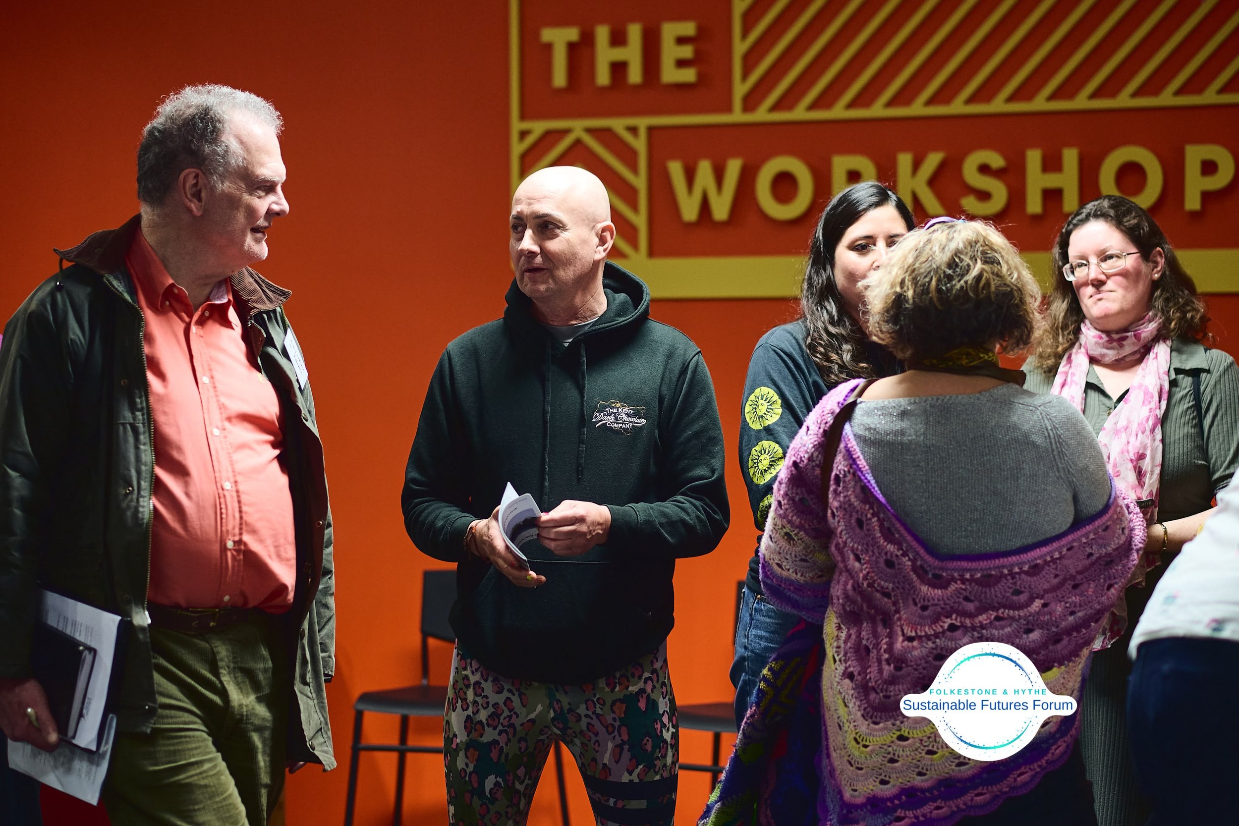 Group of five people engaged in conversation at an indoor workshop with an orange background and a sign that reads "The Workshop."