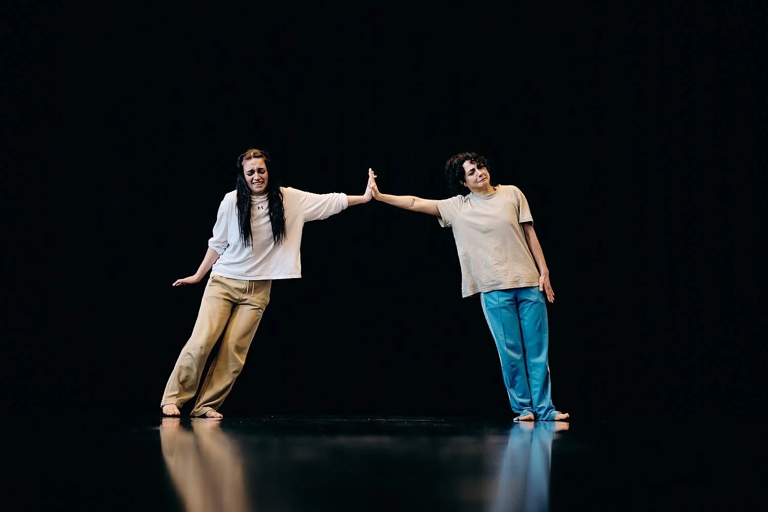 Two women performing a dance or theatrical act on a black stage, touching hands with one smiling and the other pensive, both dressed casually.