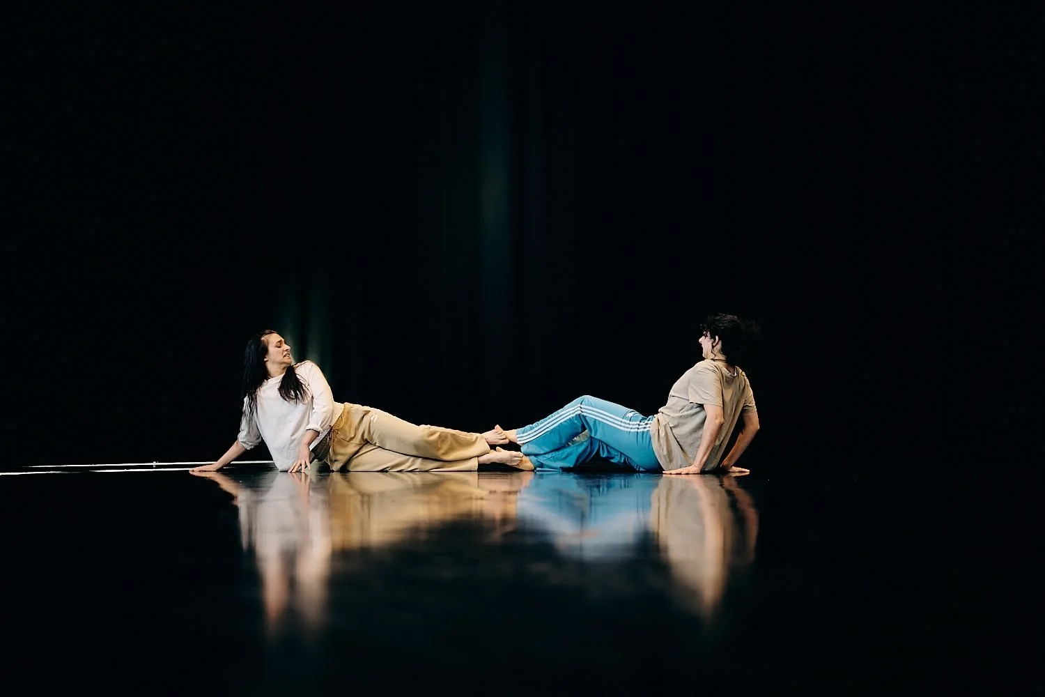 Two women are performing a dance or theatrical pose on a dark stage, holding hands with their legs extended and parallel to the floor.