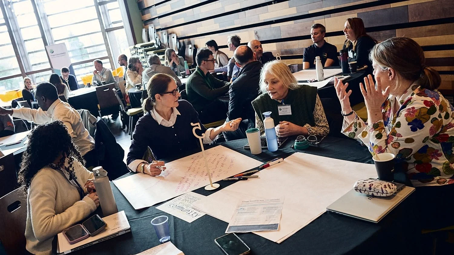 People sitting at tables in a conference room, engaging in discussions and taking notes.